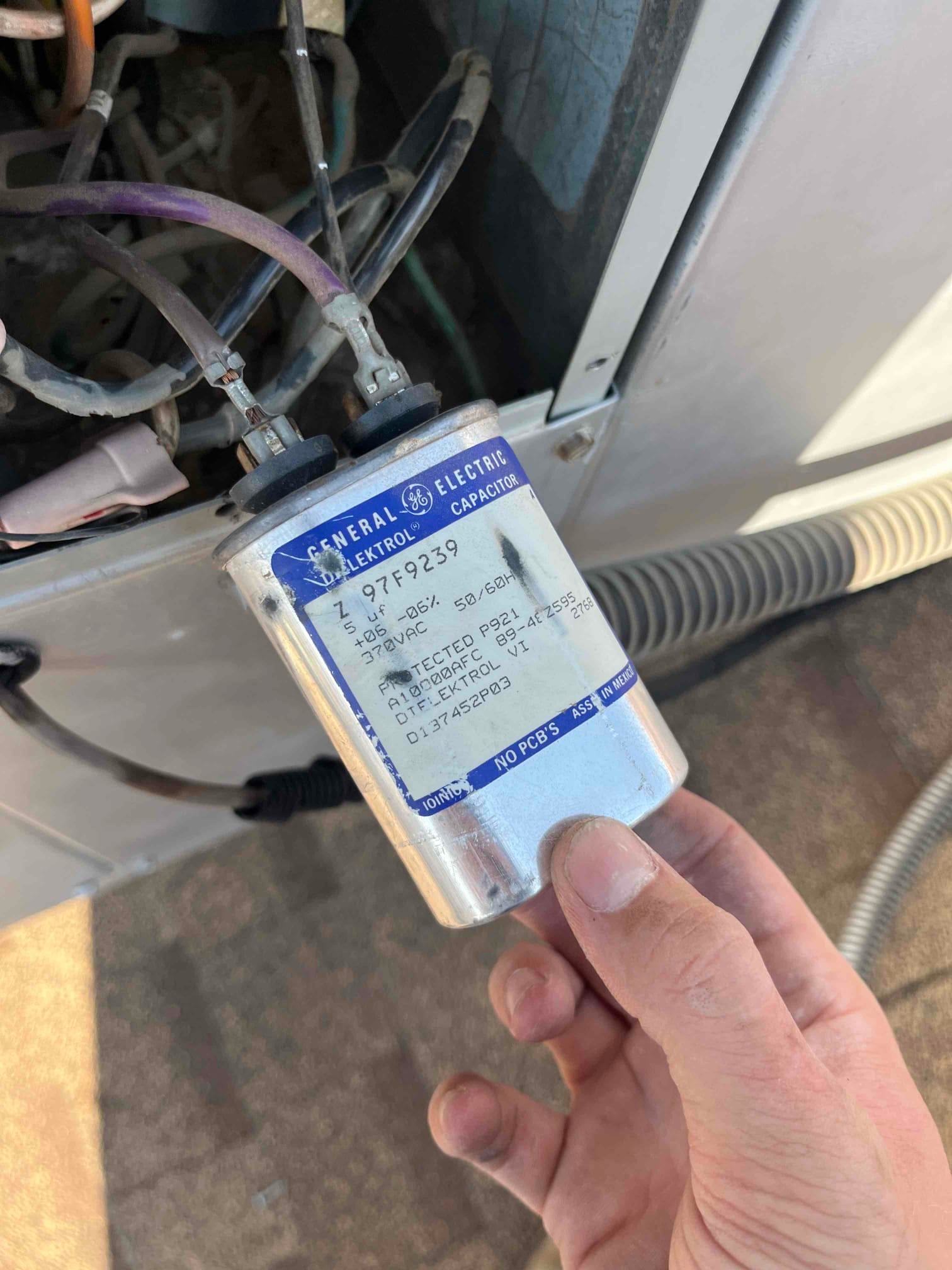 Technician holds a dual-run capacitor next to condenser wiring, showing printed rating label and clean terminals on top.