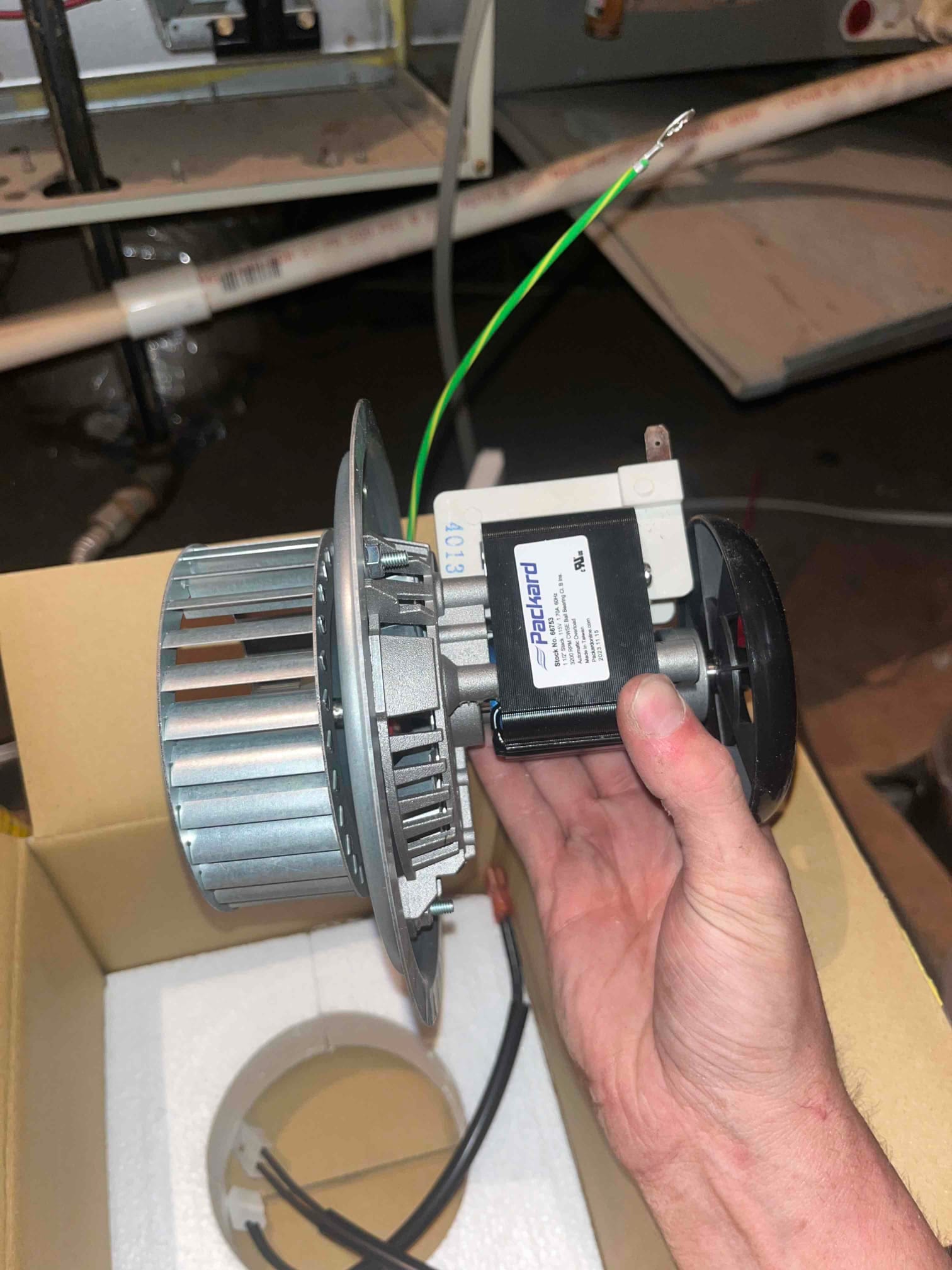 Technician holds a new furnace inducer draft motor assembly with wiring leads, showing clean housing and intact impeller.