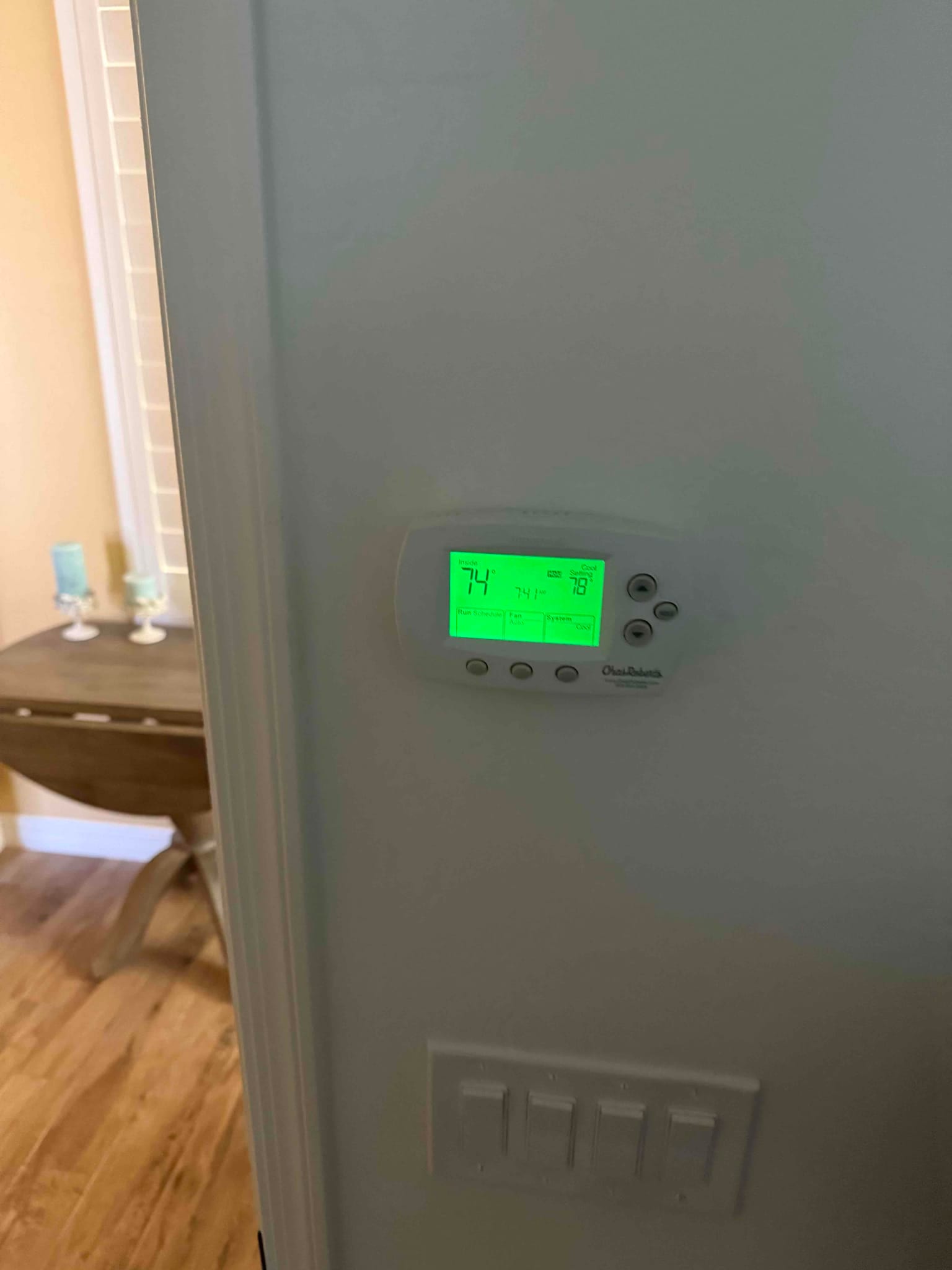 Wall-mounted digital thermostat illuminated and operating after installation, with adjacent light switches visible below on the finished wall.