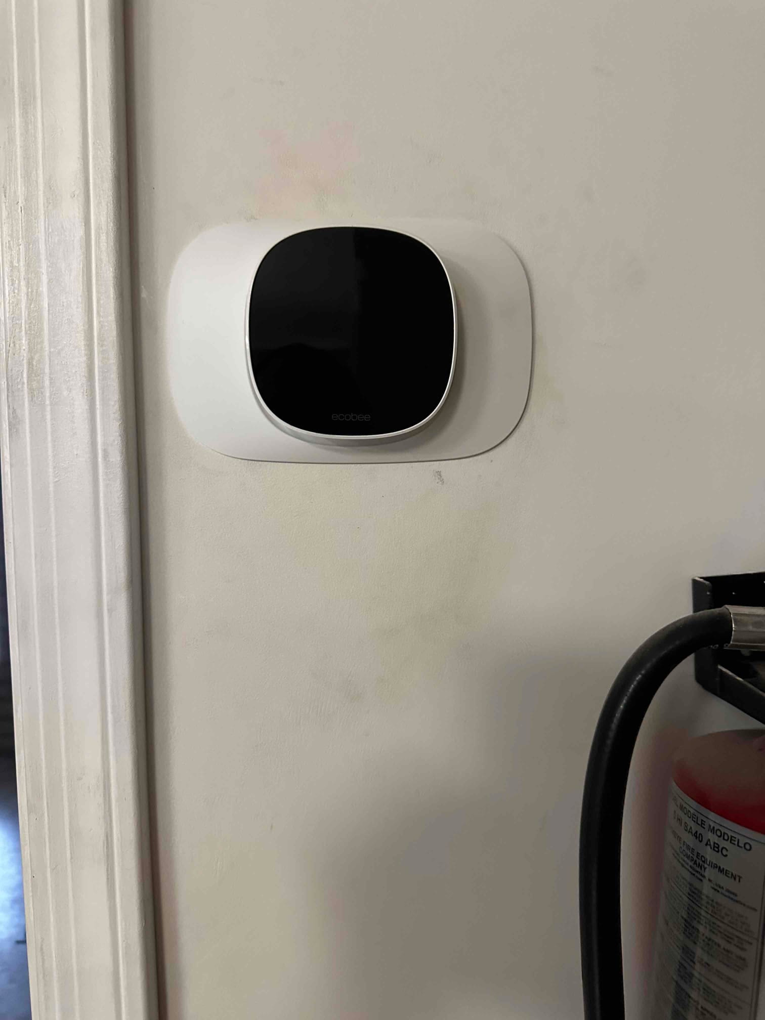 Wall-mounted smart thermostat installed with trim plate, adjacent to a fire extinguisher, as part of the HVAC control setup.