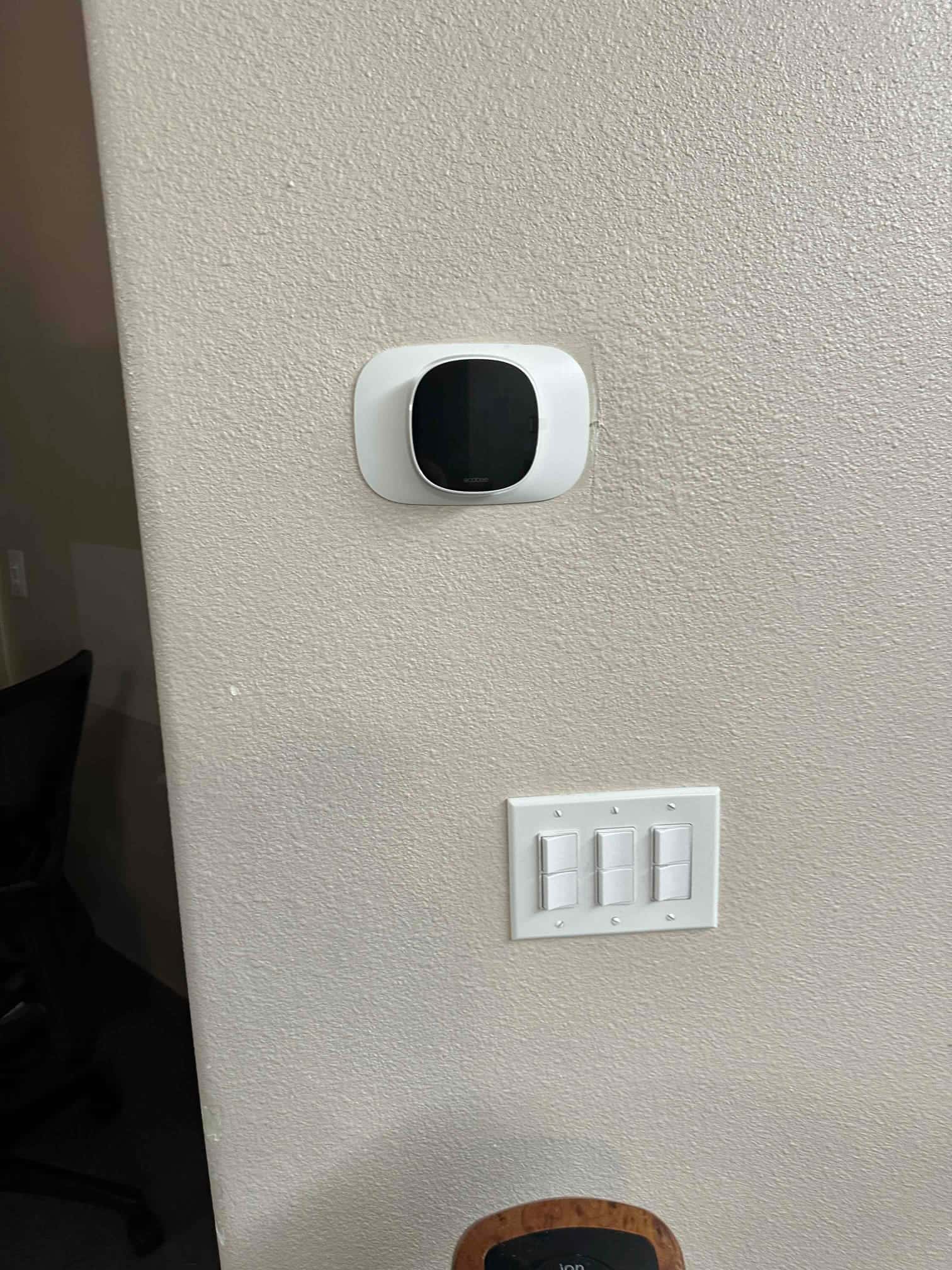 Wall-mounted smart thermostat installed with trim plate above a bank of three light switches on a finished wall.