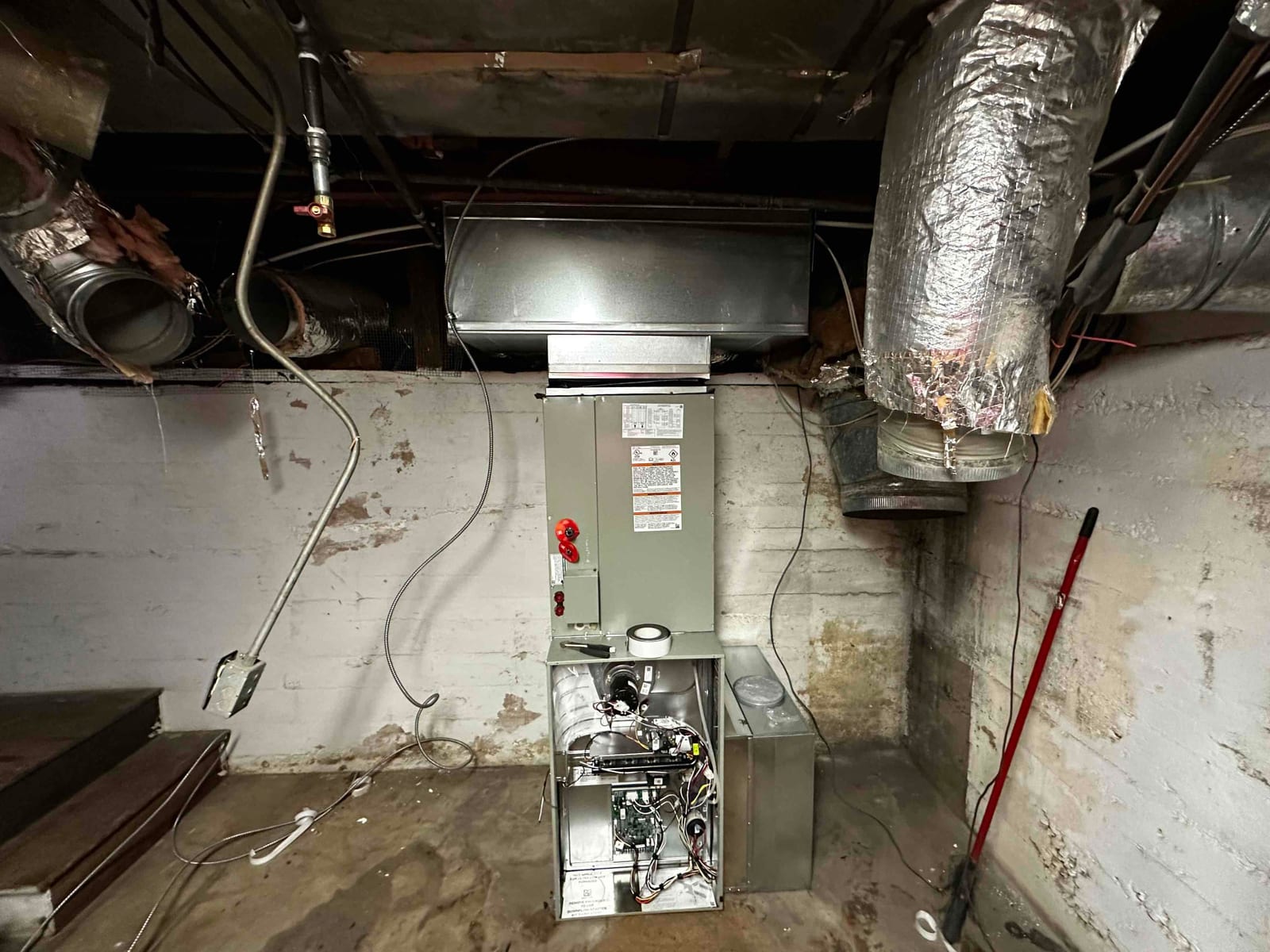 New gas furnace and duct transitions installed in a basement, with access panel open showing internal wiring and components.