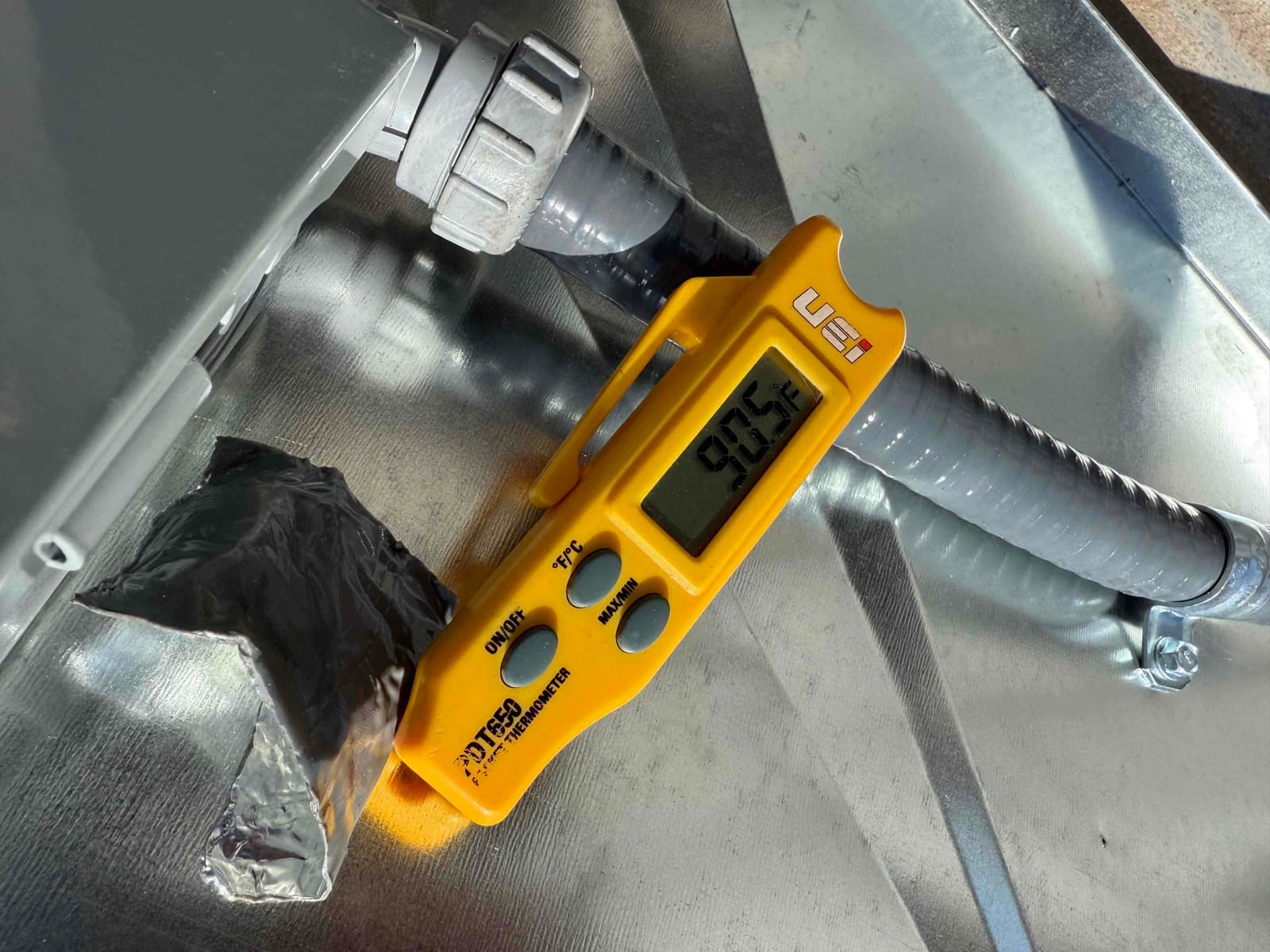 Digital thermometer placed on metal duct surface shows 90.5°F during air temperature check at the new installation.