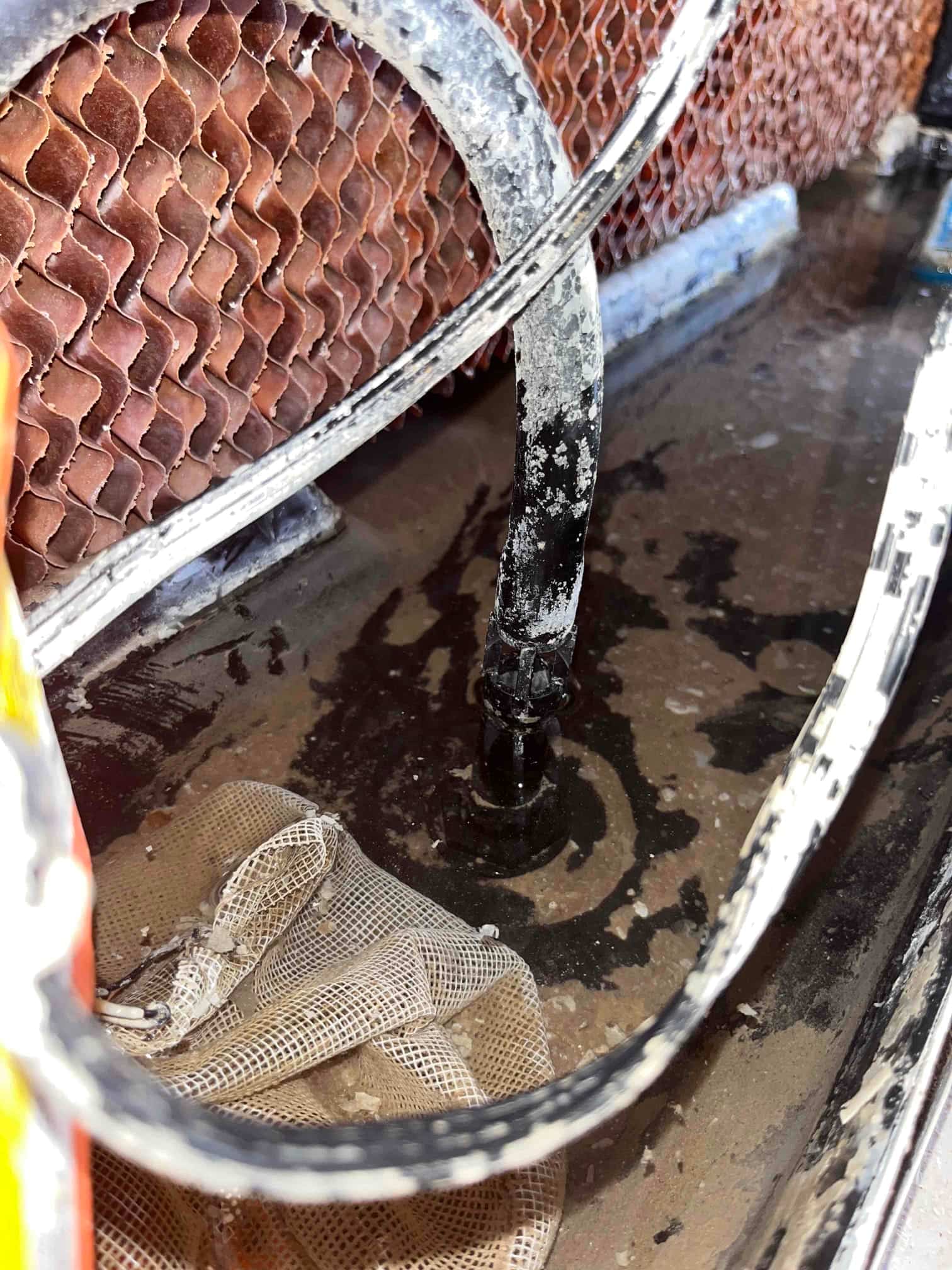 Evaporative cooler sump area showing the stand pipe connection, wet basin surface, and adjacent mesh debris screen.