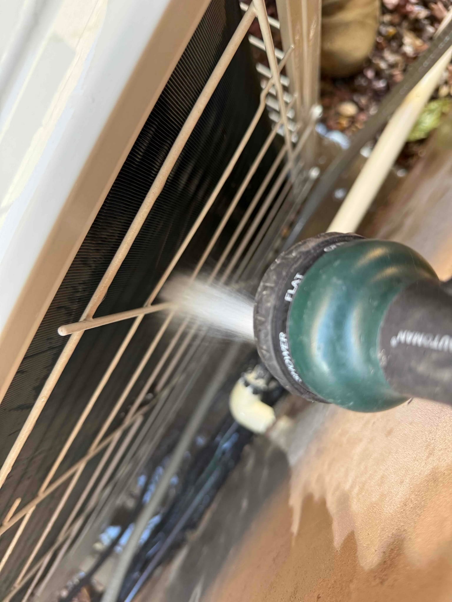 Garden hose nozzle sprays water through the grille to wash debris from the outdoor condenser coil fins.