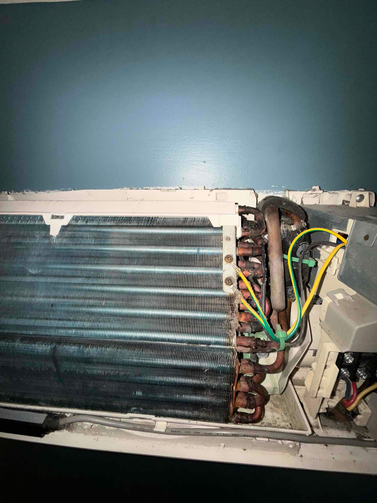 Mini-split indoor evaporator coil exposed, showing copper tubing, finned coil surface, and connected wiring before detailed cleaning.