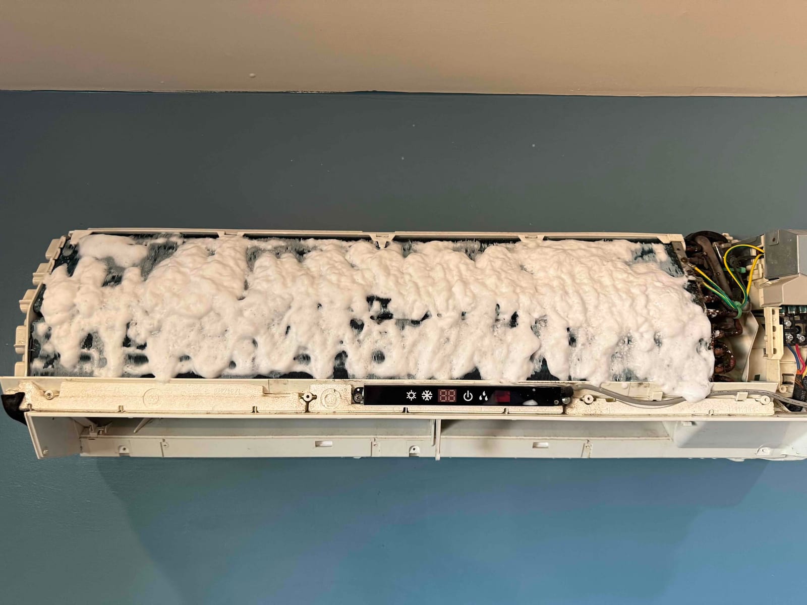Mini-split indoor evaporator coil foams with cleaner during coil cleaning, with cover removed to expose fins and electronics.