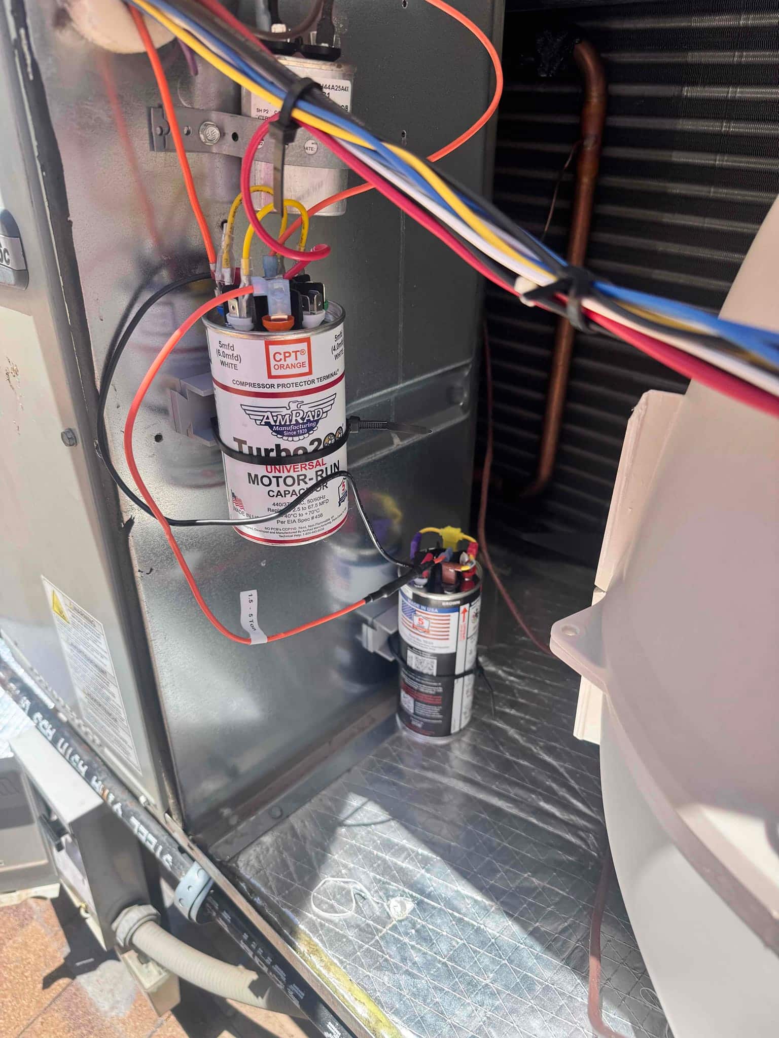 New motor run capacitor installed with connected wiring, and an additional capacitor mounted nearby inside the condenser unit compartment.