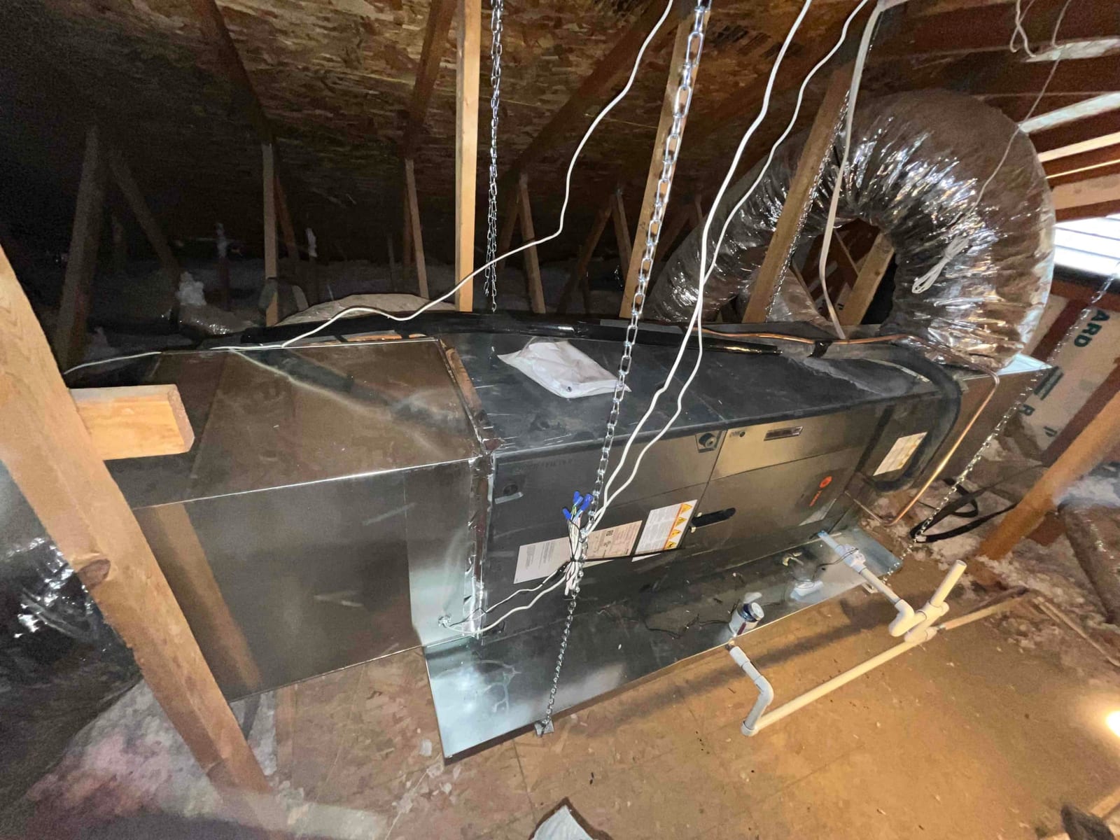 Air handler suspended in attic with insulated duct connections, new sheet-metal plenum, and condensate drain piping installed on a service pan.
