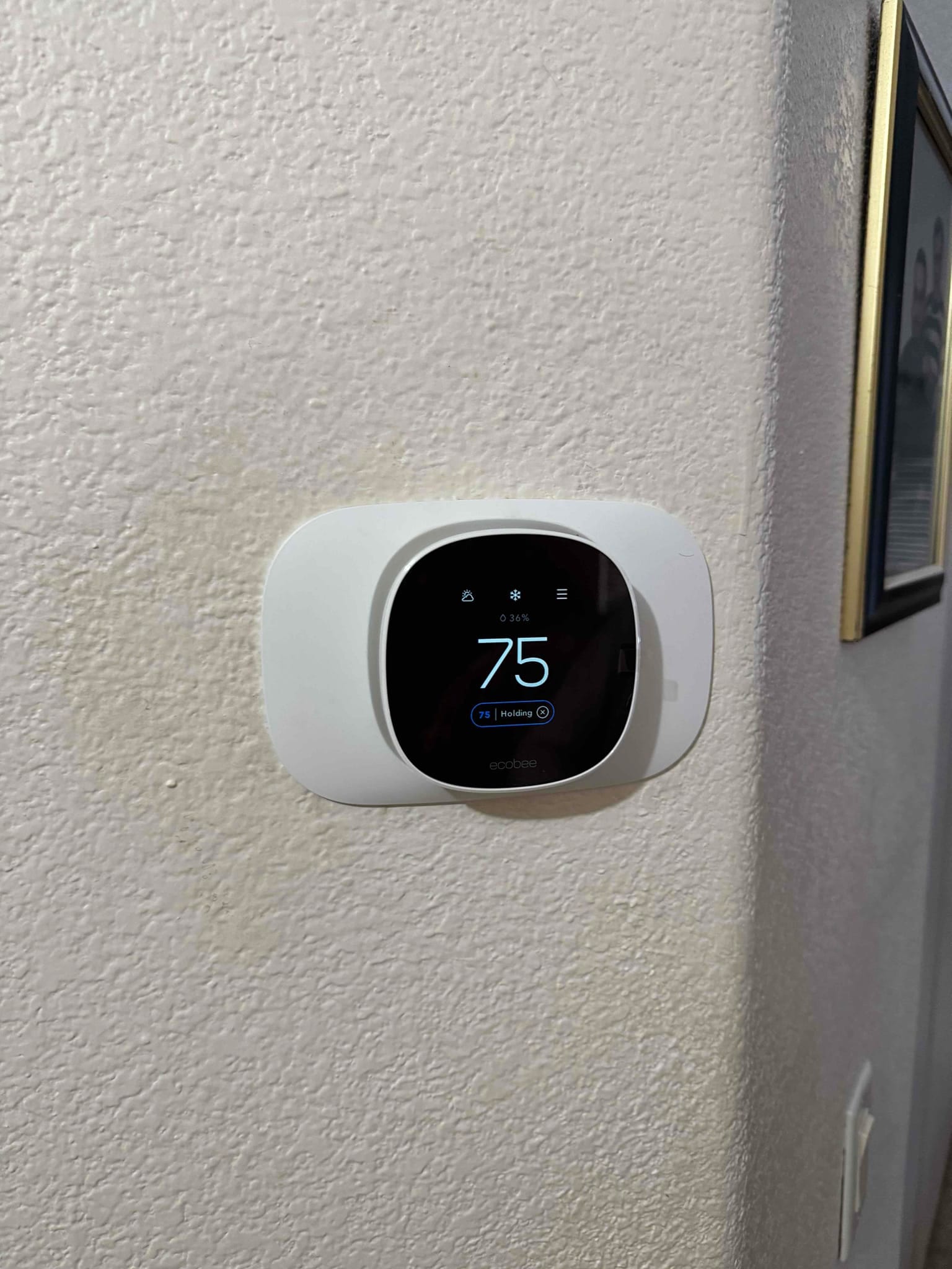 New smart thermostat installed on interior wall, display shows 75 degrees with cooling status and touch controls visible.