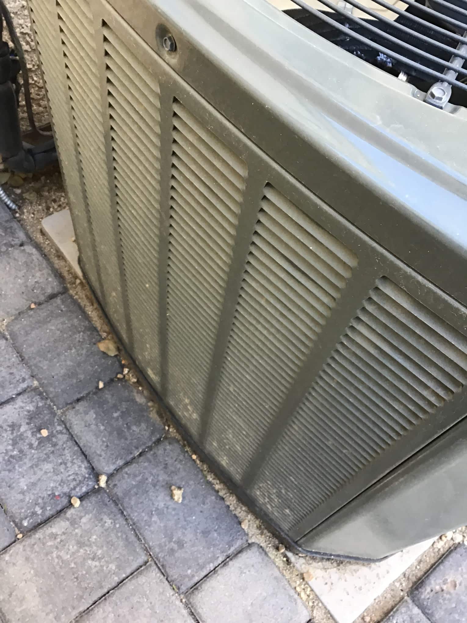 Outdoor condenser cabinet with louvered panels and surface dust, installed beside paver walkway.
