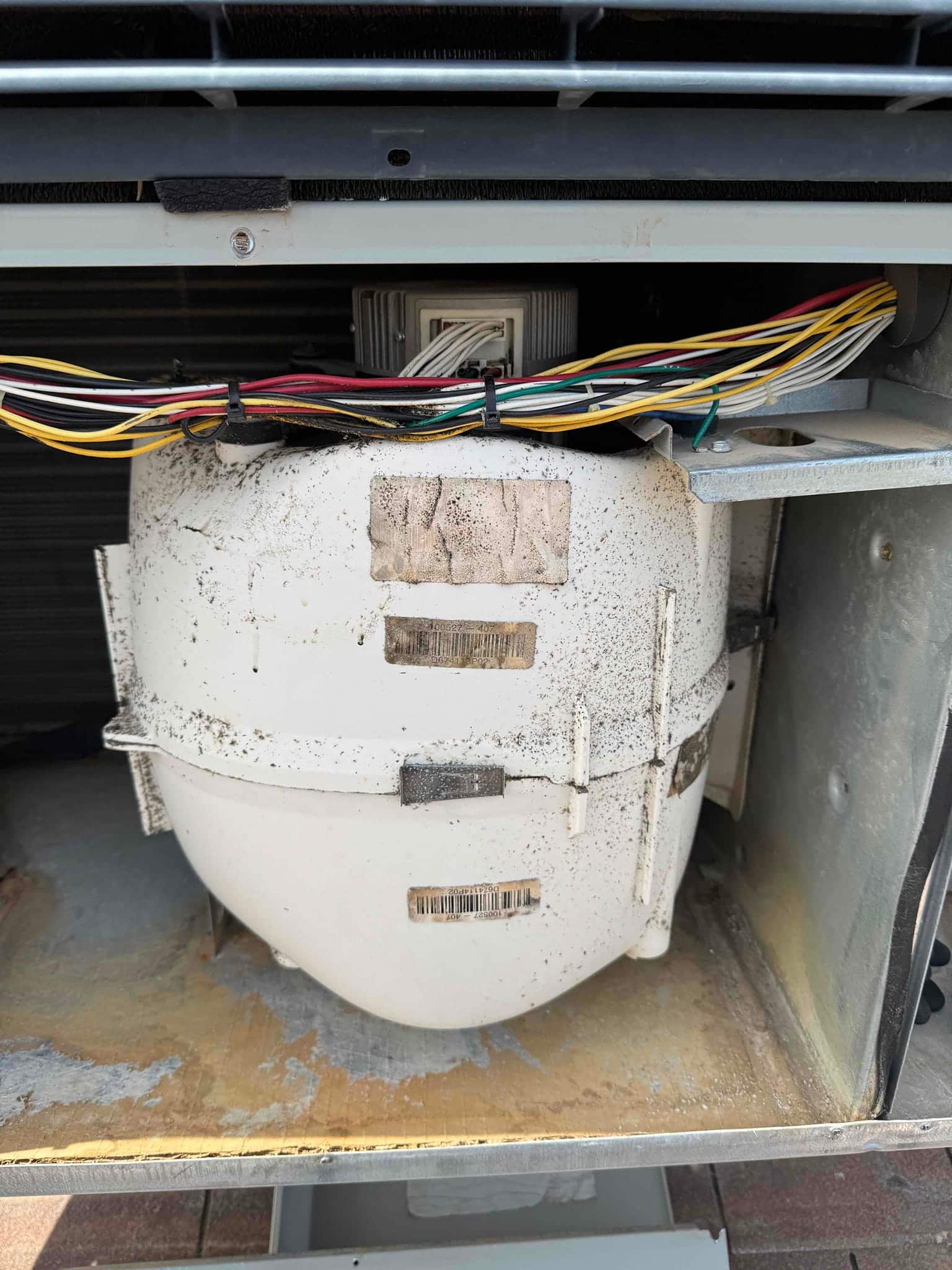 Outdoor condenser compressor housing shown with surface rust, dirt accumulation, and bundled control wiring above the unit.