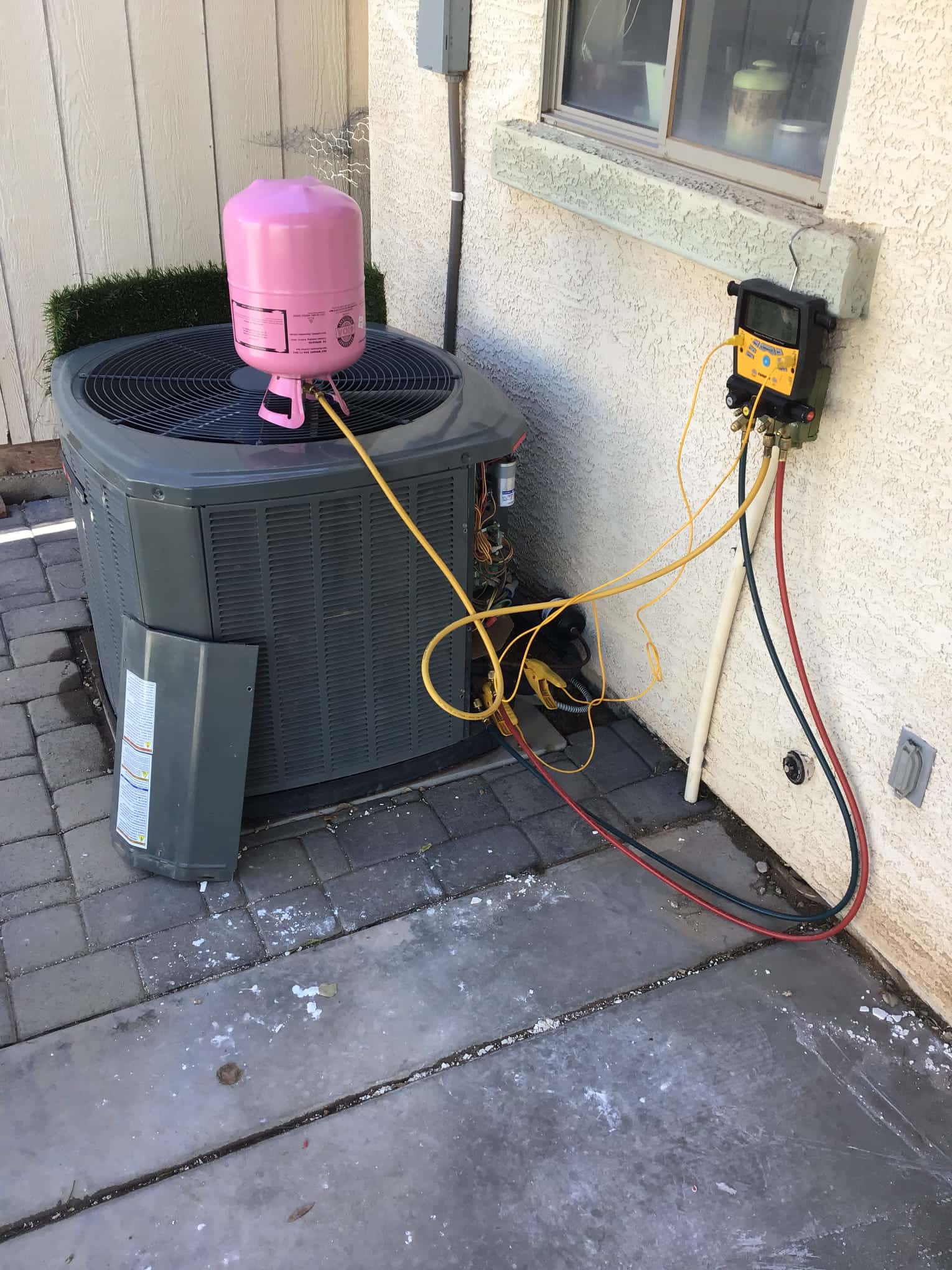 Outdoor condenser with service panel open, digital manifold mounted on wall, hoses connected to unit, and pink refrigerant cylinder on top.