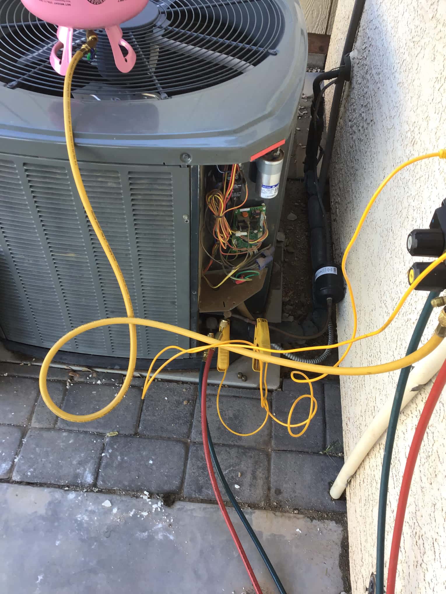 Outdoor condenser with service panel open showing wiring and a mounted run capacitor, connected to refrigerant hoses and a cylinder during setup.