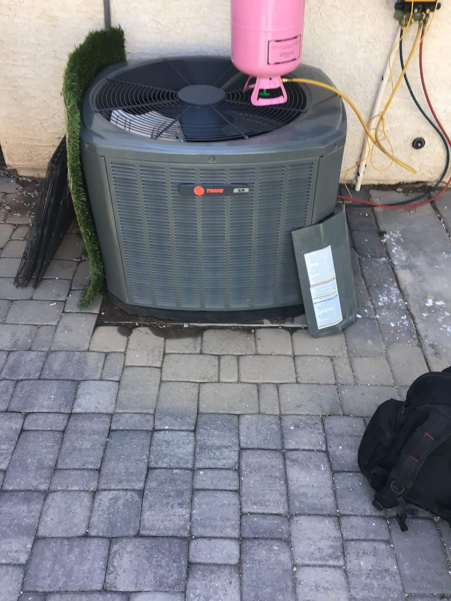 Outdoor condenser with side access panel removed; refrigerant cylinder on top with yellow hose connected to service line.