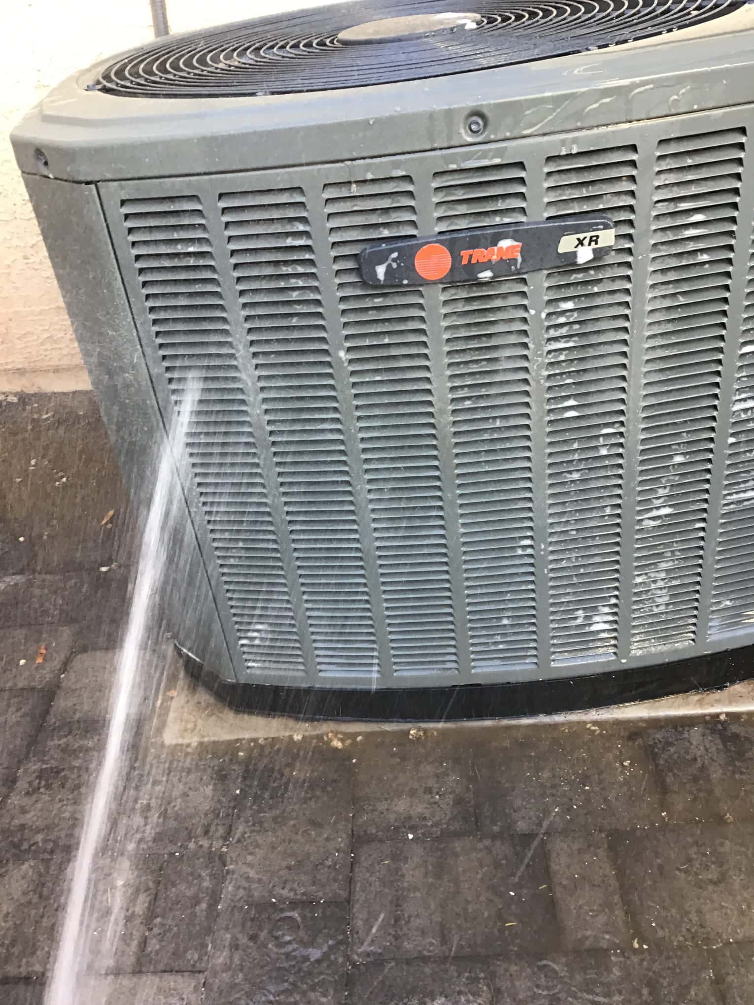 Outdoor condenser unit with front grille and top fan shroud, being rinsed with a water spray.