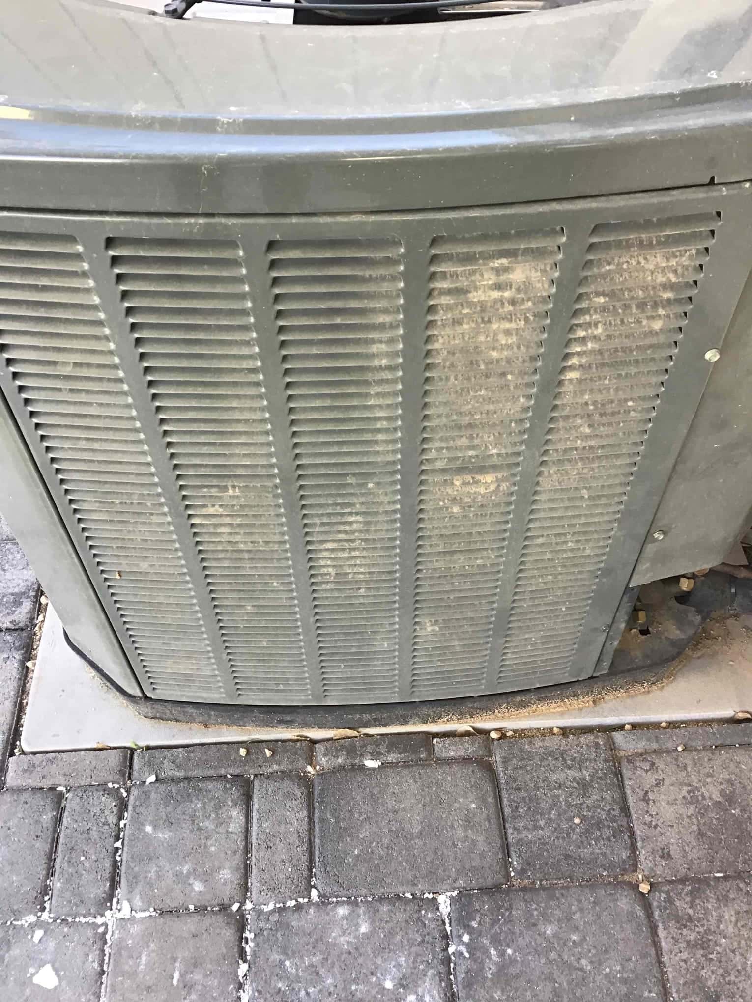 Outdoor condenser unit with louvered grille; dust and debris visible on cabinet surface and base.