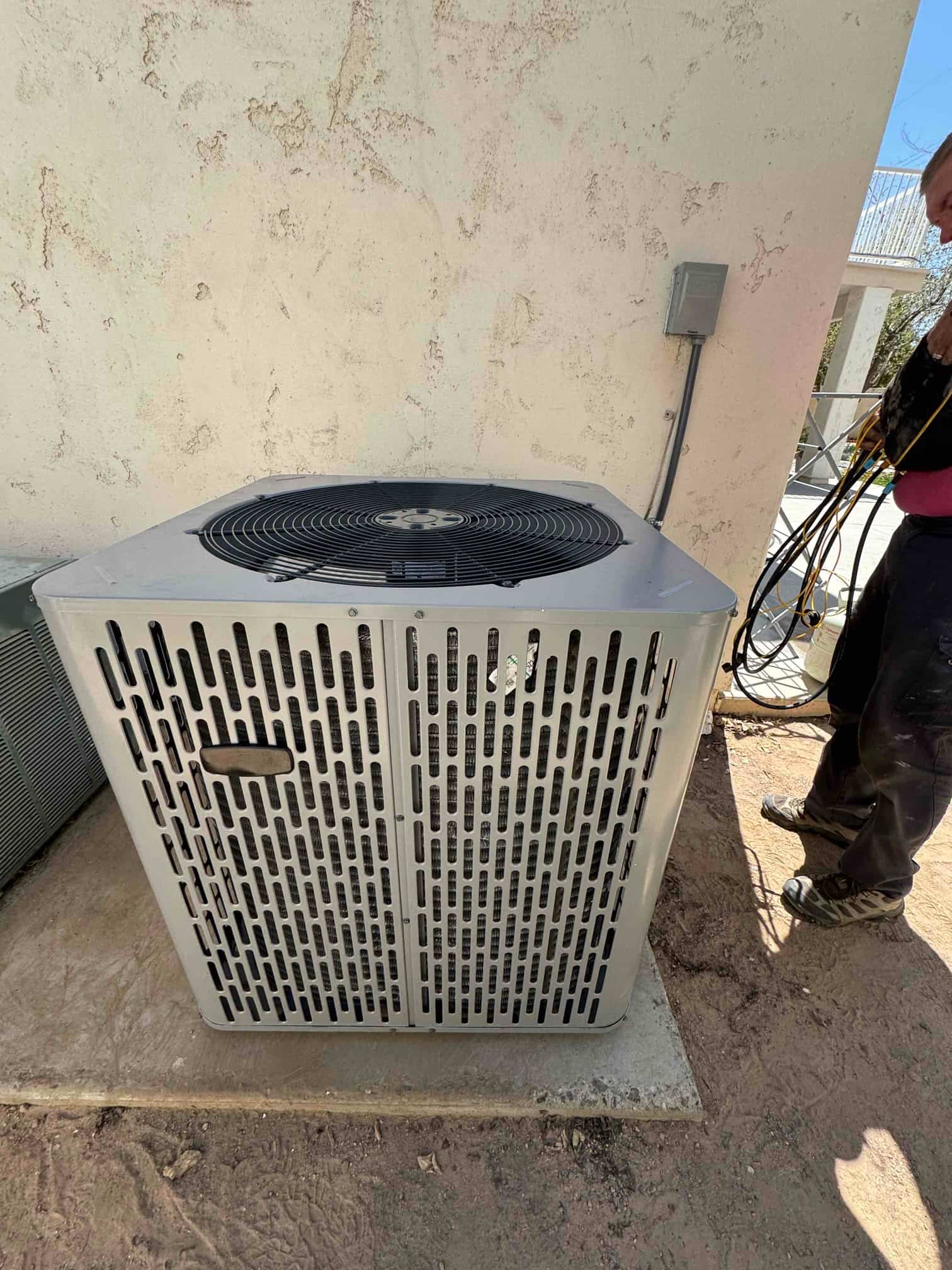 Outdoor condenser set on a level pad with electrical disconnect mounted on the wall and refrigerant hoses nearby.