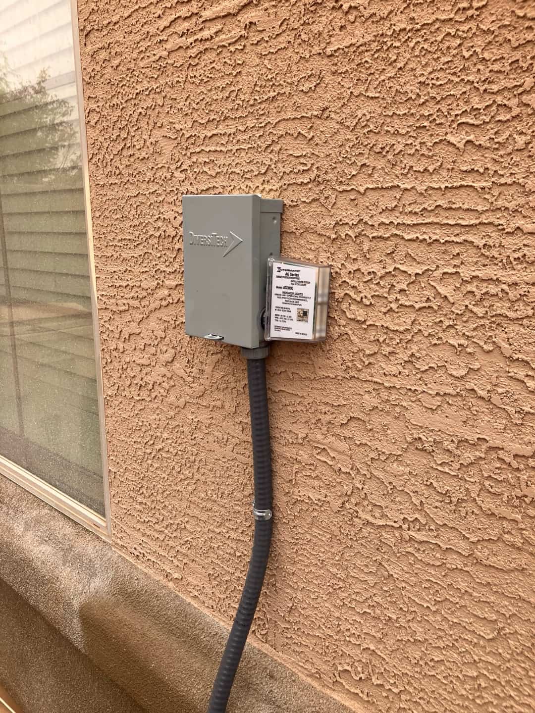Outdoor electrical disconnect with flexible conduit installed on exterior wall beside window, providing service power for the condenser unit.