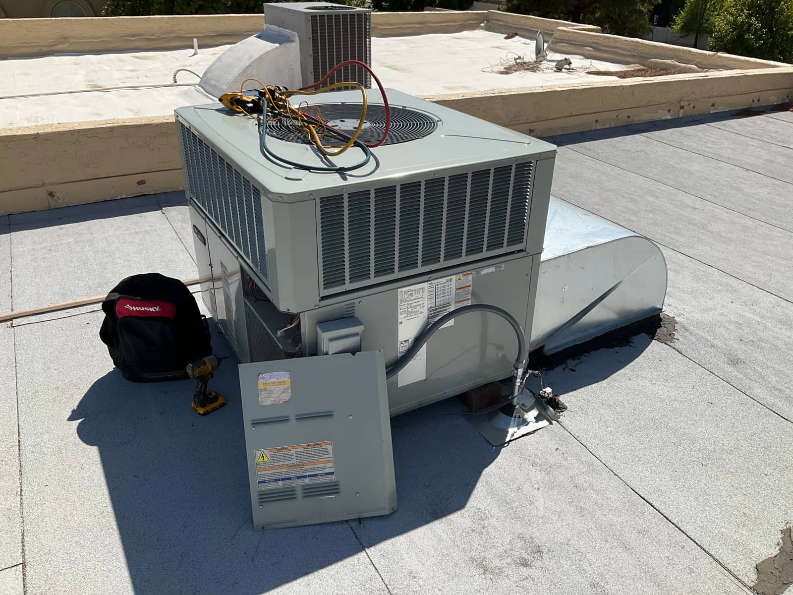 Rooftop package HVAC unit with service panel open; manifold hoses and clamp meter rest on top during inspection.