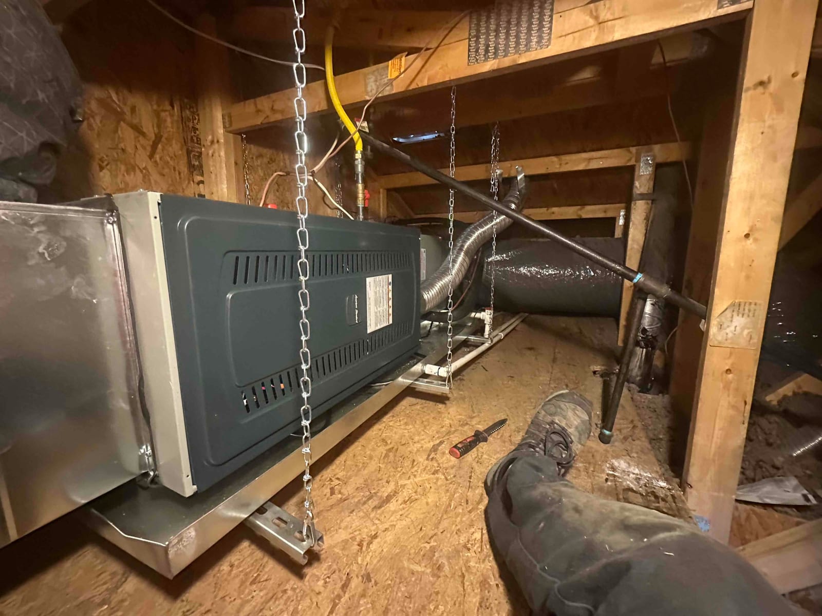 Suspended air handler installed on hangers with drain pan, flexible duct connections, gas line, and tools visible in the attic.
