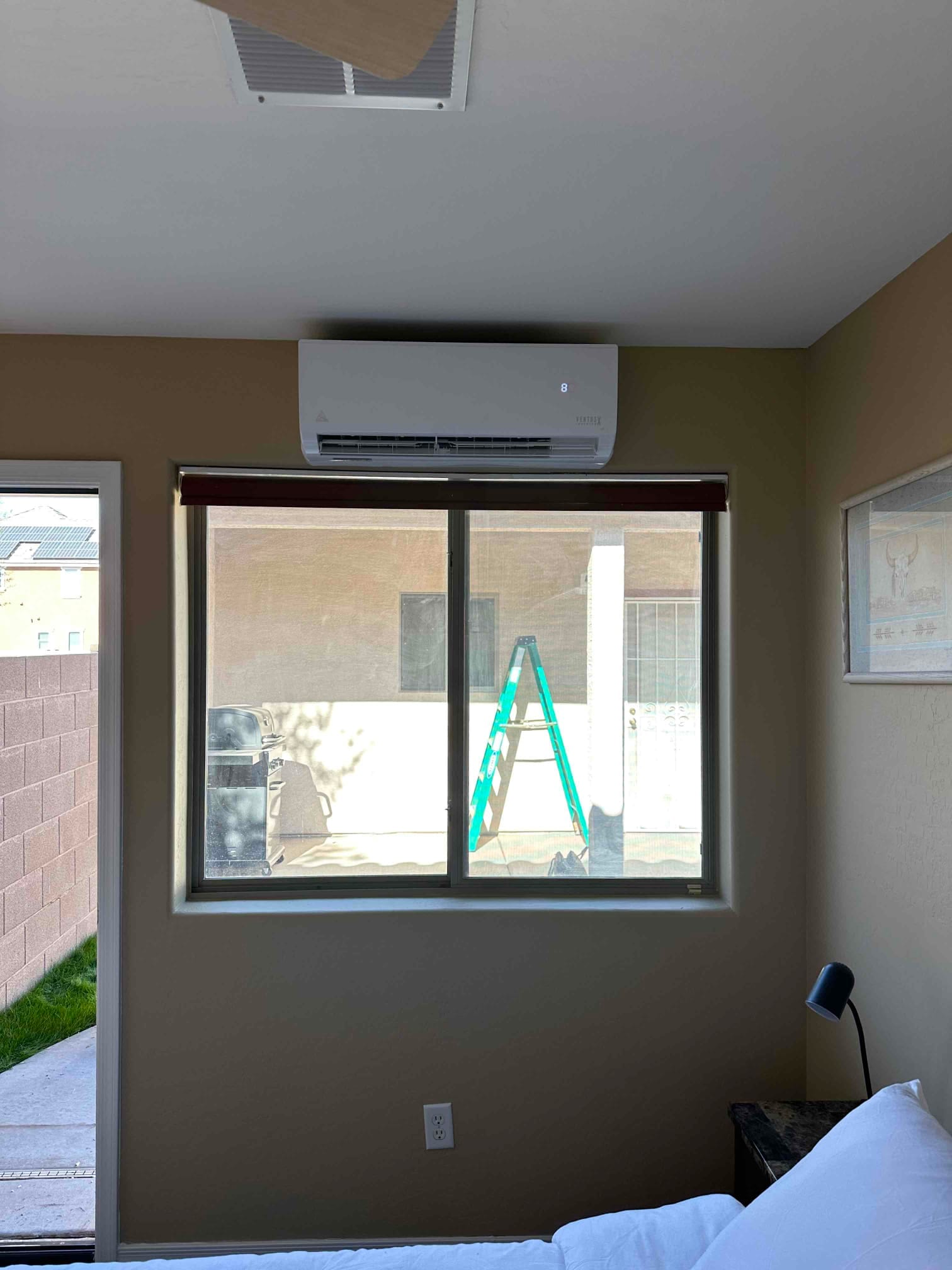Wall-mounted ductless indoor unit installed above a window, powered on, with finished wall penetration and nearby supply grille on ceiling.