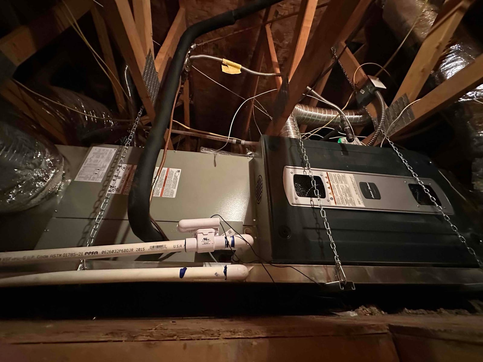 New attic air handler and furnace assembly suspended on chains with insulated refrigerant line and PVC condensate drain installed.
