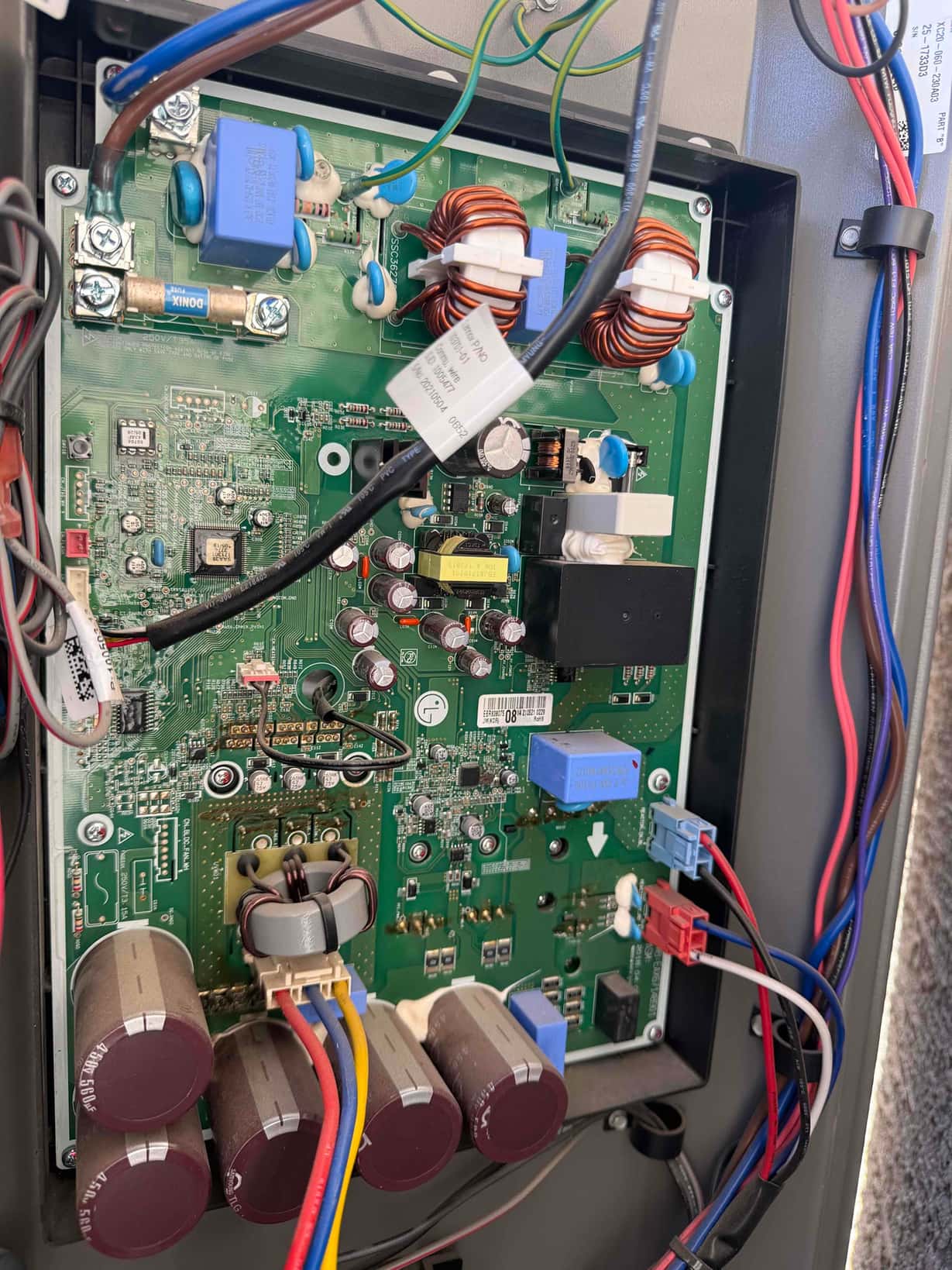 Outdoor inverter control board exposed during repair, showing wiring connections, transformers, relays, and multiple large capacitors.