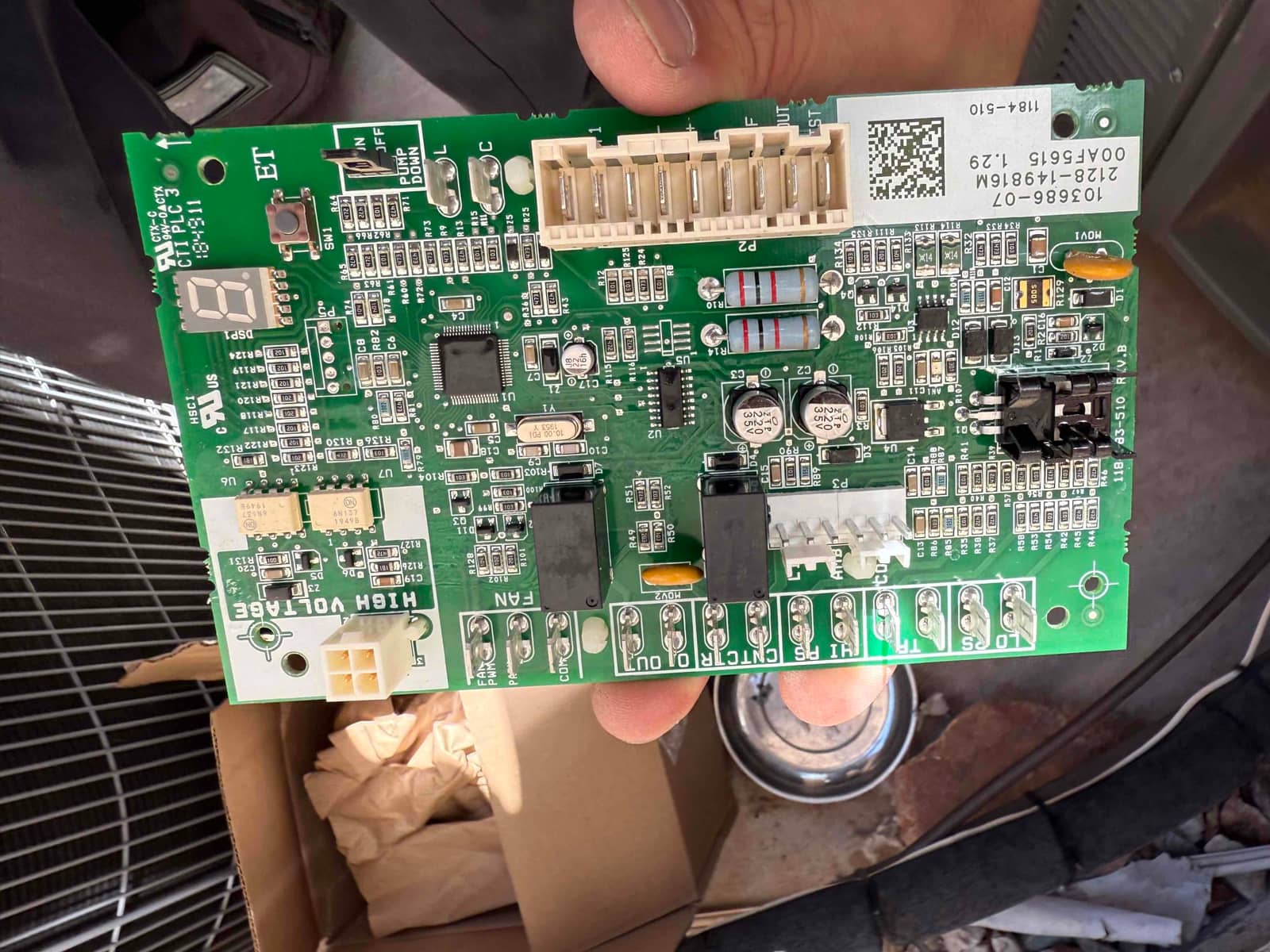 Technician holds an outdoor unit control board showing connectors, relays, fuses, and labeled terminals for fan and high voltage.