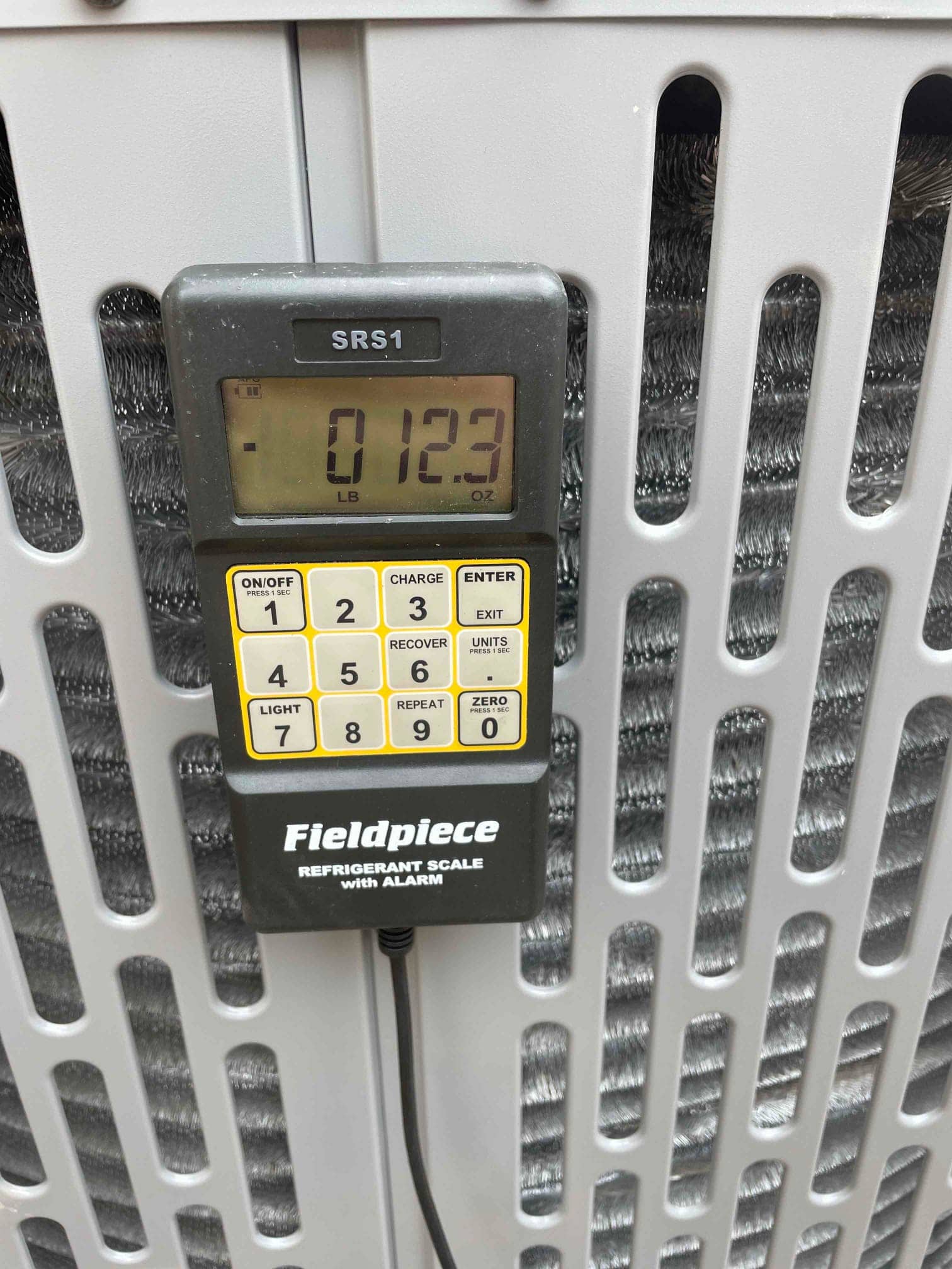 Digital refrigerant scale hanging on the condenser cabinet shows a reading of 0 lb 12.3 oz during charging.