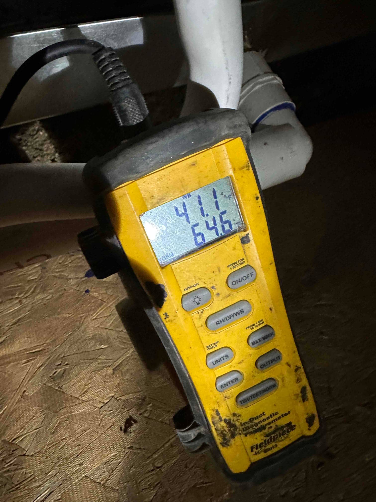Digital thermometer shows 64.6°F air temperature during system airflow check near the indoor unit drain piping.