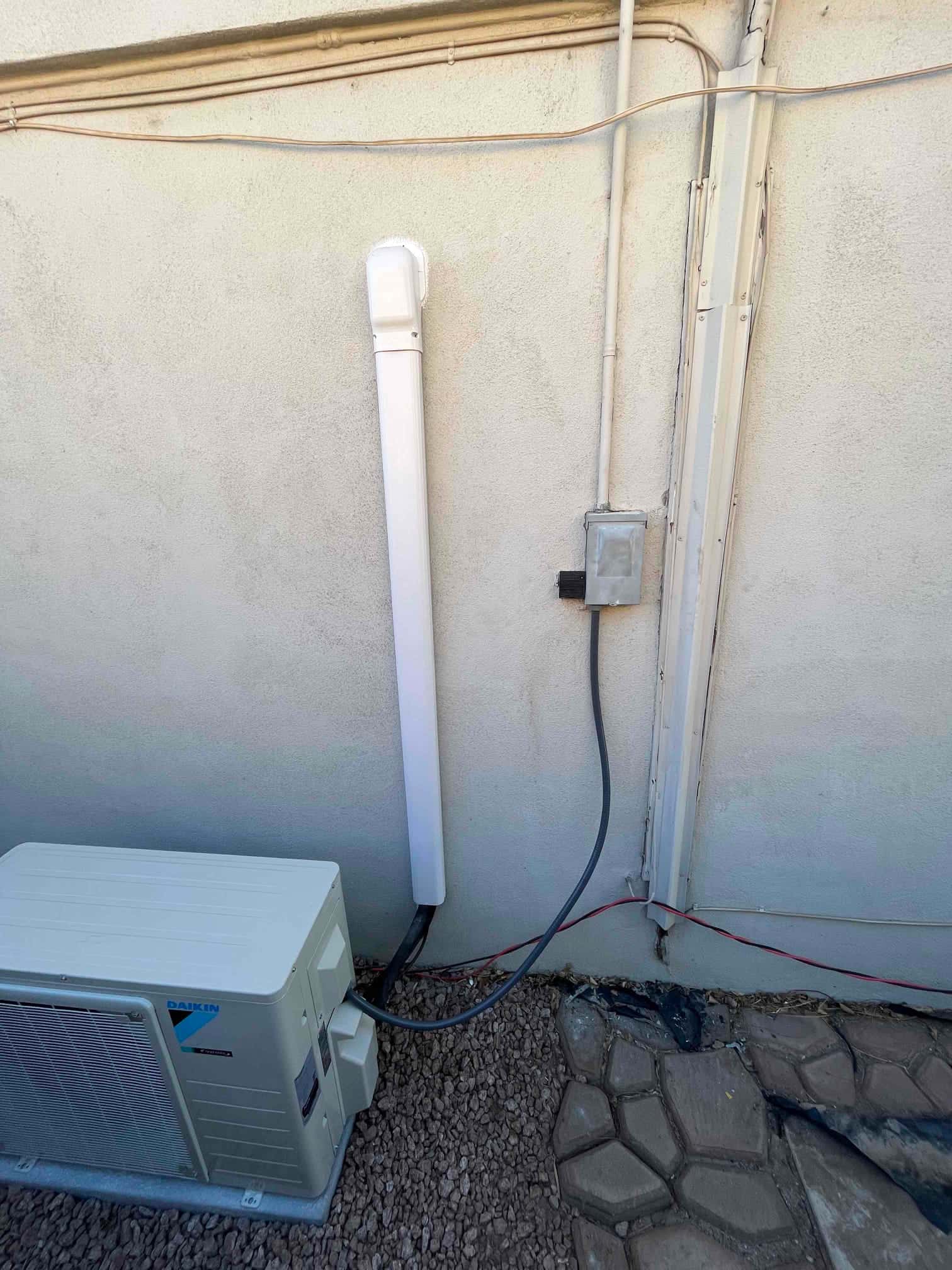 Outdoor mini-split condenser set on a pad with line-set cover, electrical disconnect, and routed conduits along the exterior wall.