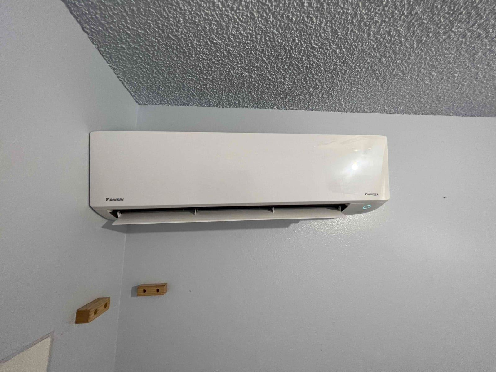 Wall-mounted ductless indoor unit installed and operating, with discharge louver open and indicator light illuminated against a finished wall.