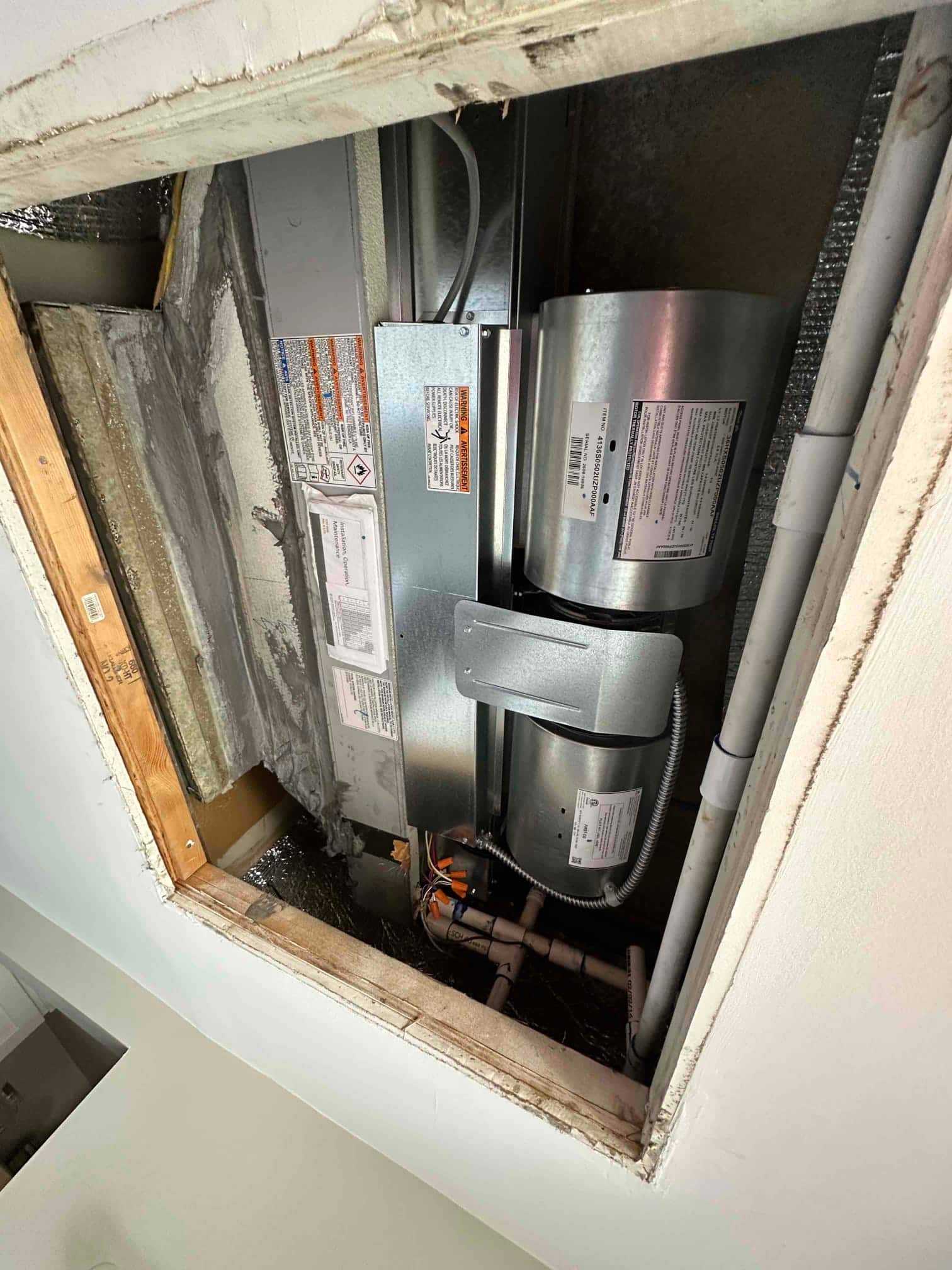 Ceiling-mounted air handler installed with insulated duct connections, electrical whip, and PVC condensate drain assembly visible in the access opening.