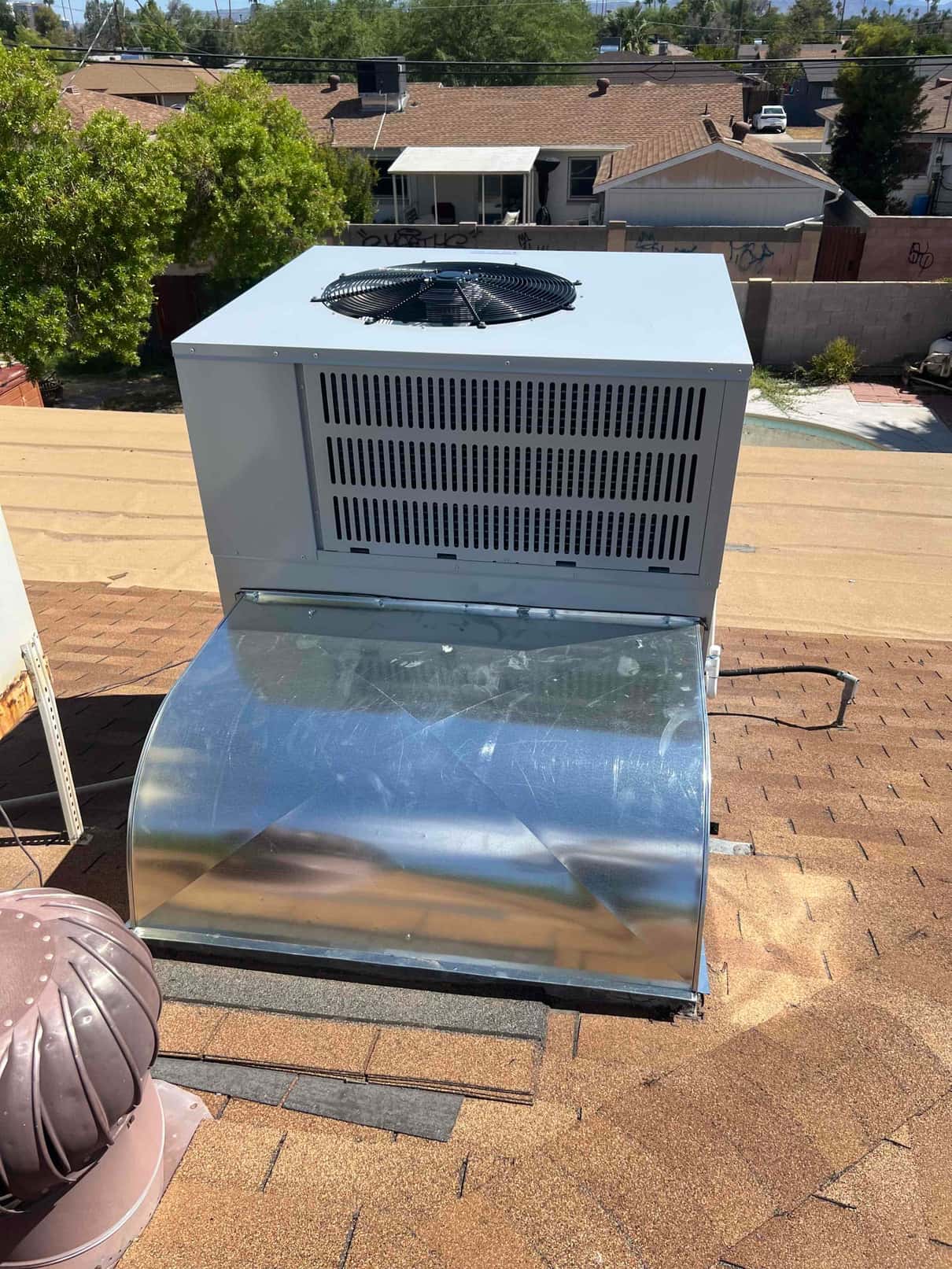 New rooftop packaged heat pump set on curb with sheet metal transition duct connected, viewed from above on shingle roof.