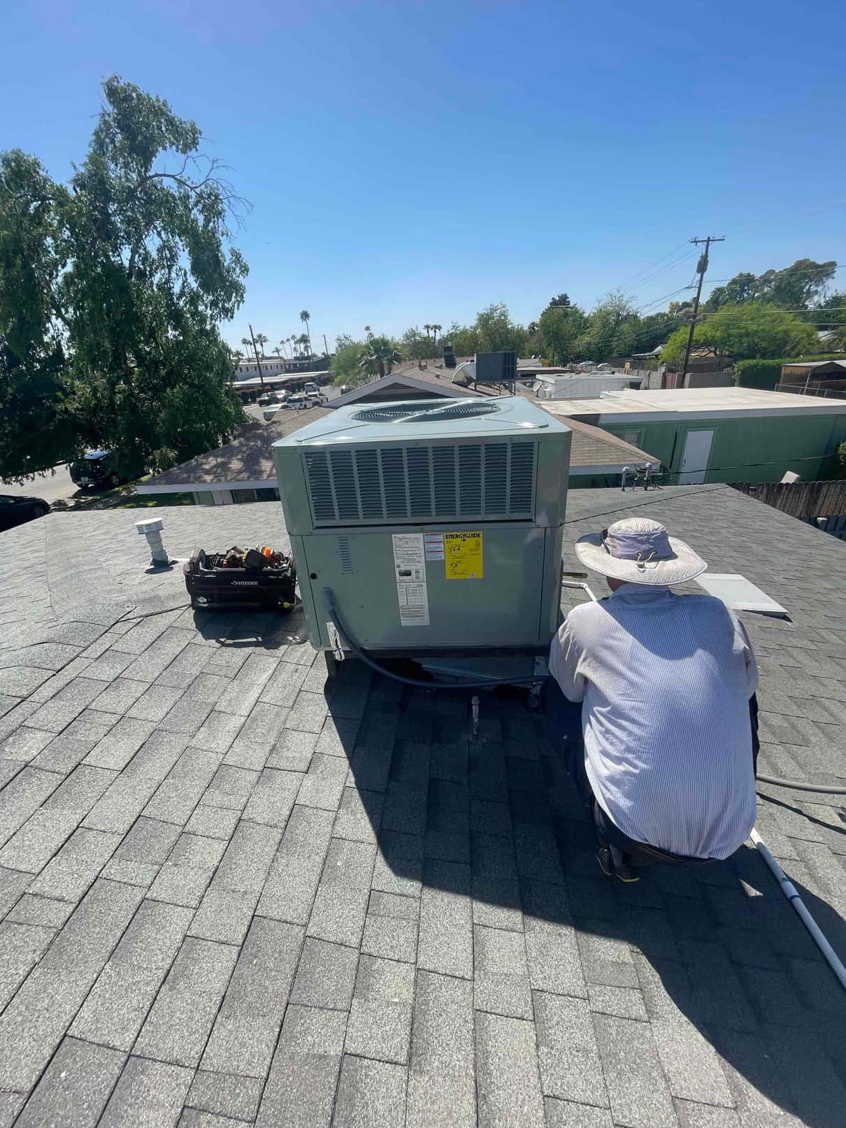 Rooftop package HVAC unit installed on shingles with technician working at the service panel and tools nearby.