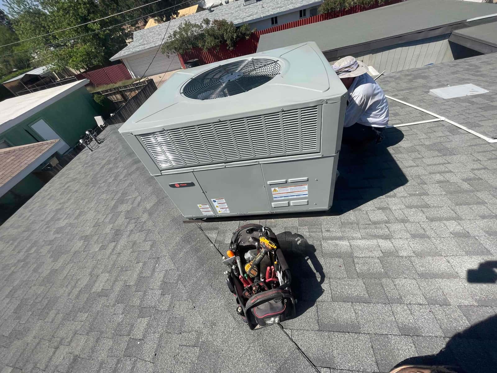 Rooftop packaged HVAC unit set on shingles with technician working at the access panel and tools staged nearby.