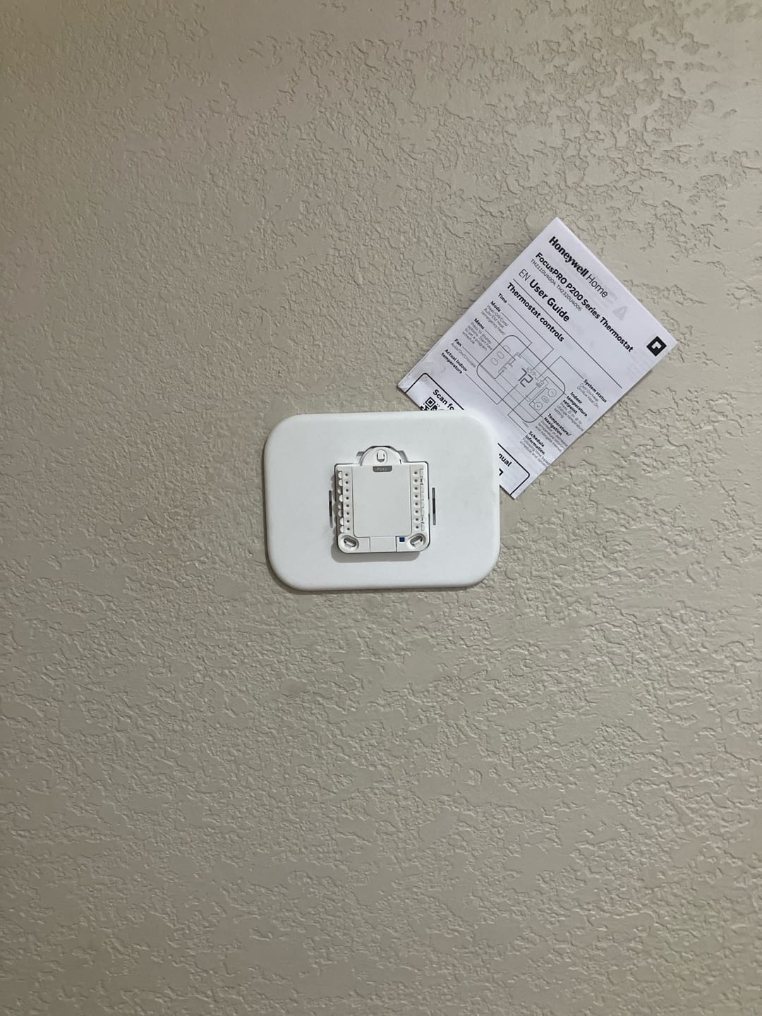 Thermostat wall plate and mounting base installed on interior wall with user guide taped nearby for reference.