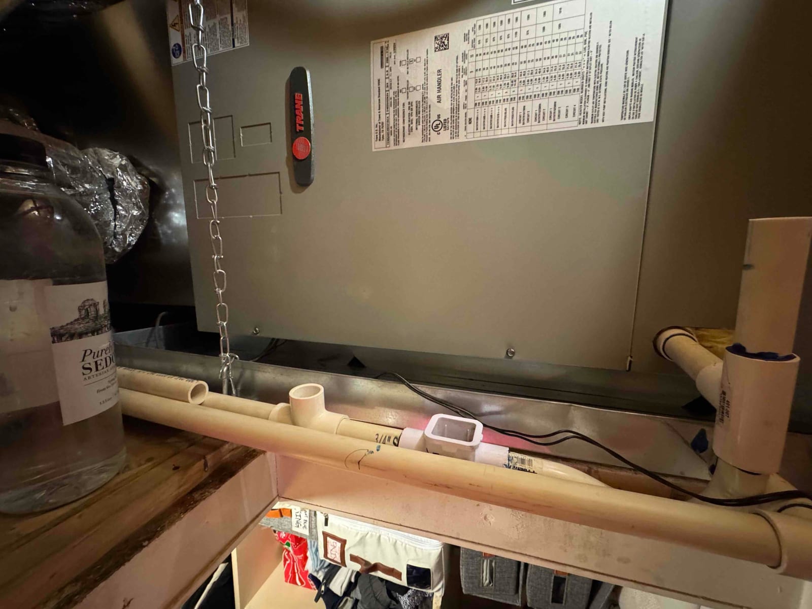 Condensate drain line assembly beneath an air handler, with multiple PVC fittings and cleanout tees visible.