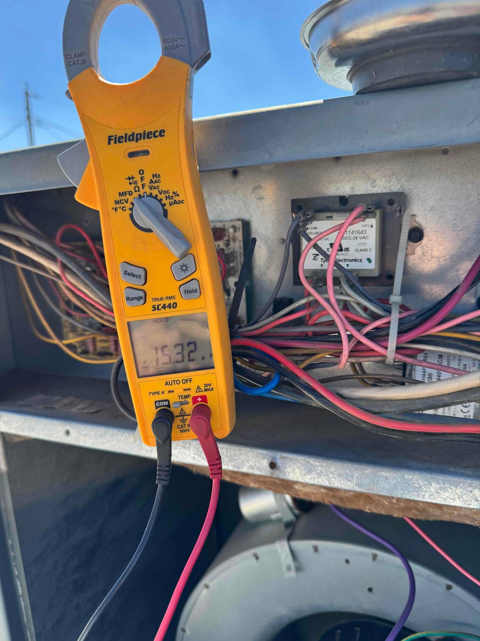 Digital multimeter displays 15.32 V while probed across two low-voltage control terminals inside the outdoor condenser panel.