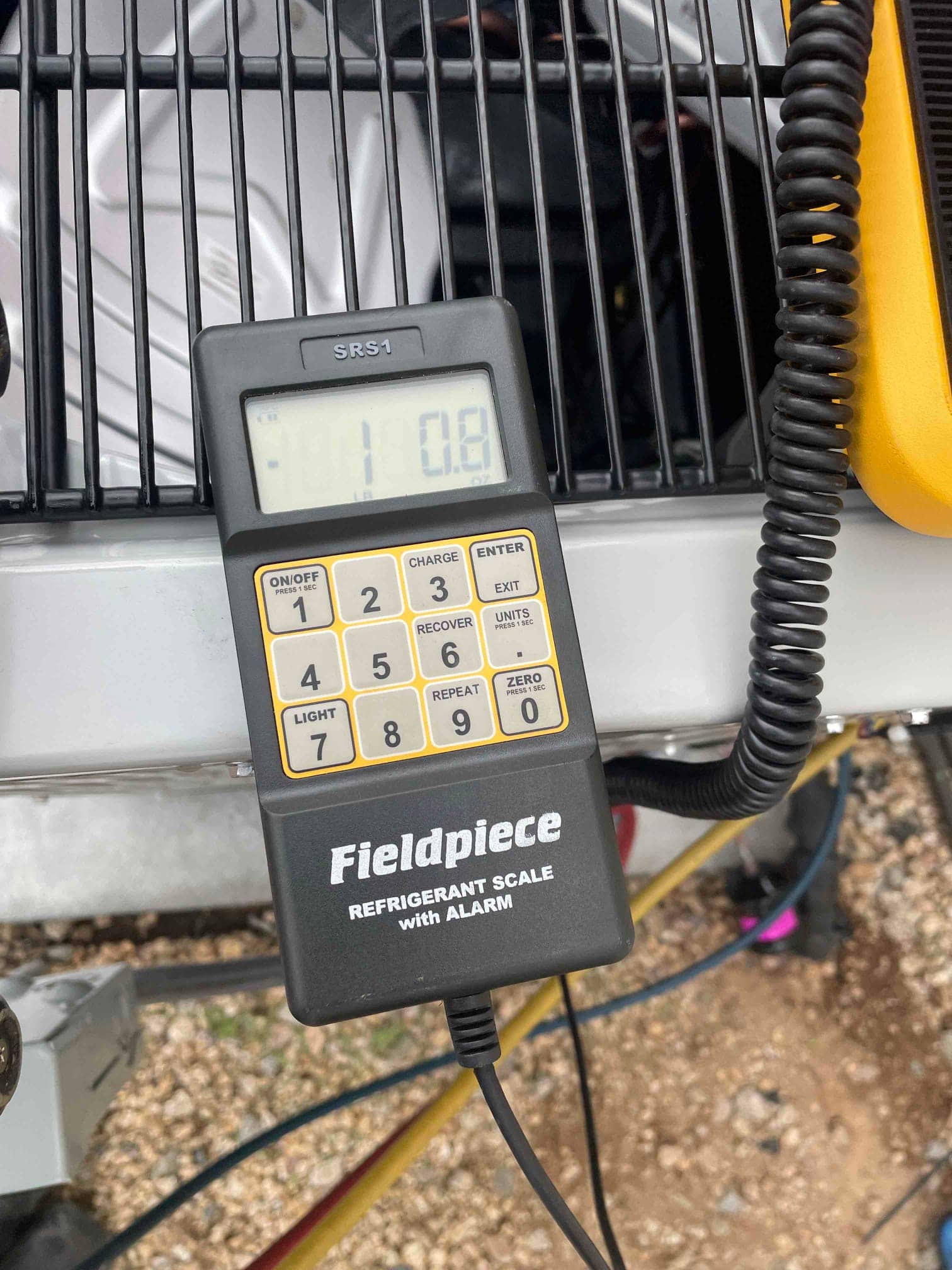 Digital refrigerant charging scale controller rests on the condenser top while connected, displaying a partial numeric reading.
