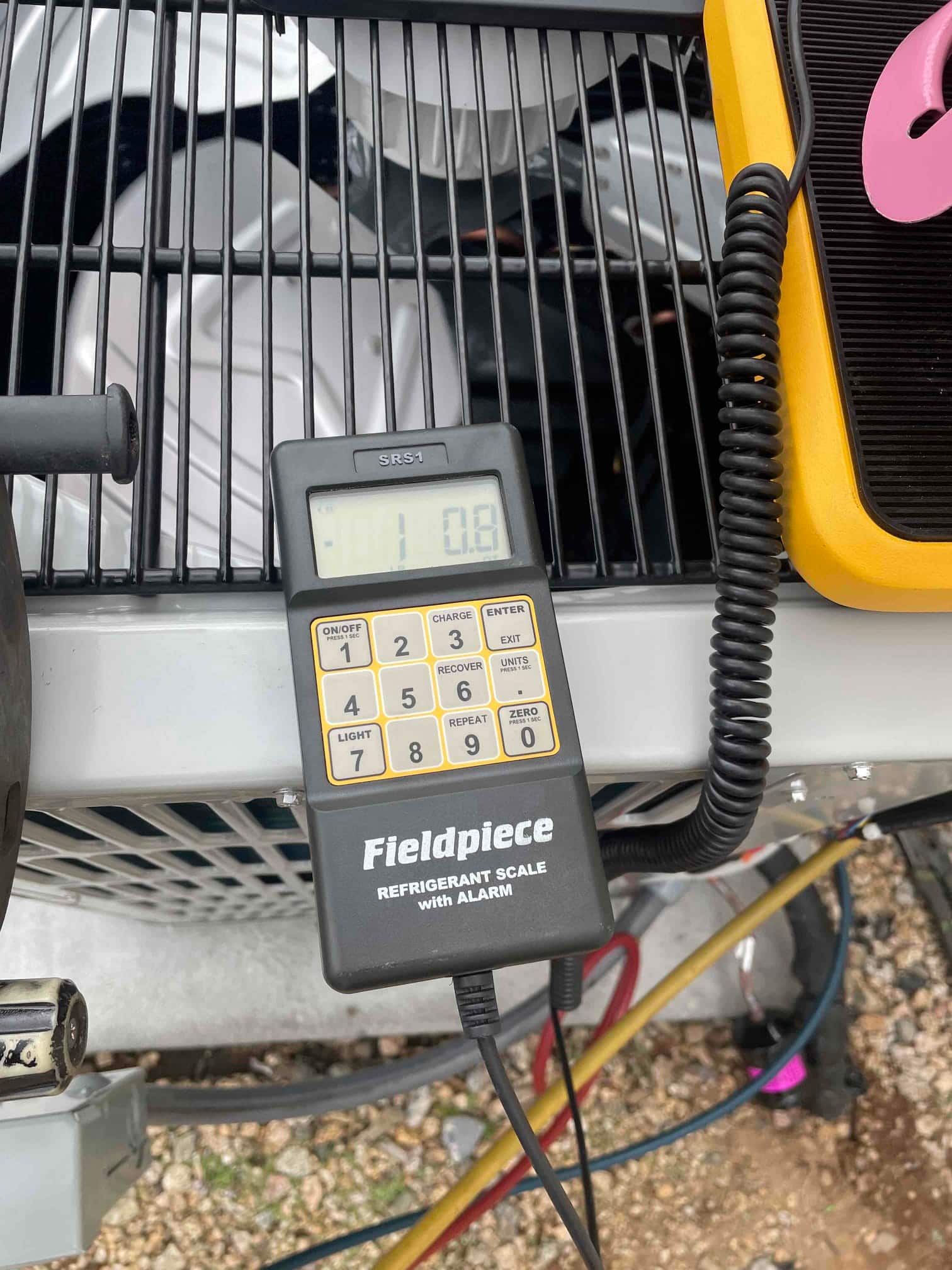 Digital refrigerant scale connected during charging beside the outdoor condenser, with hoses routed to the system.