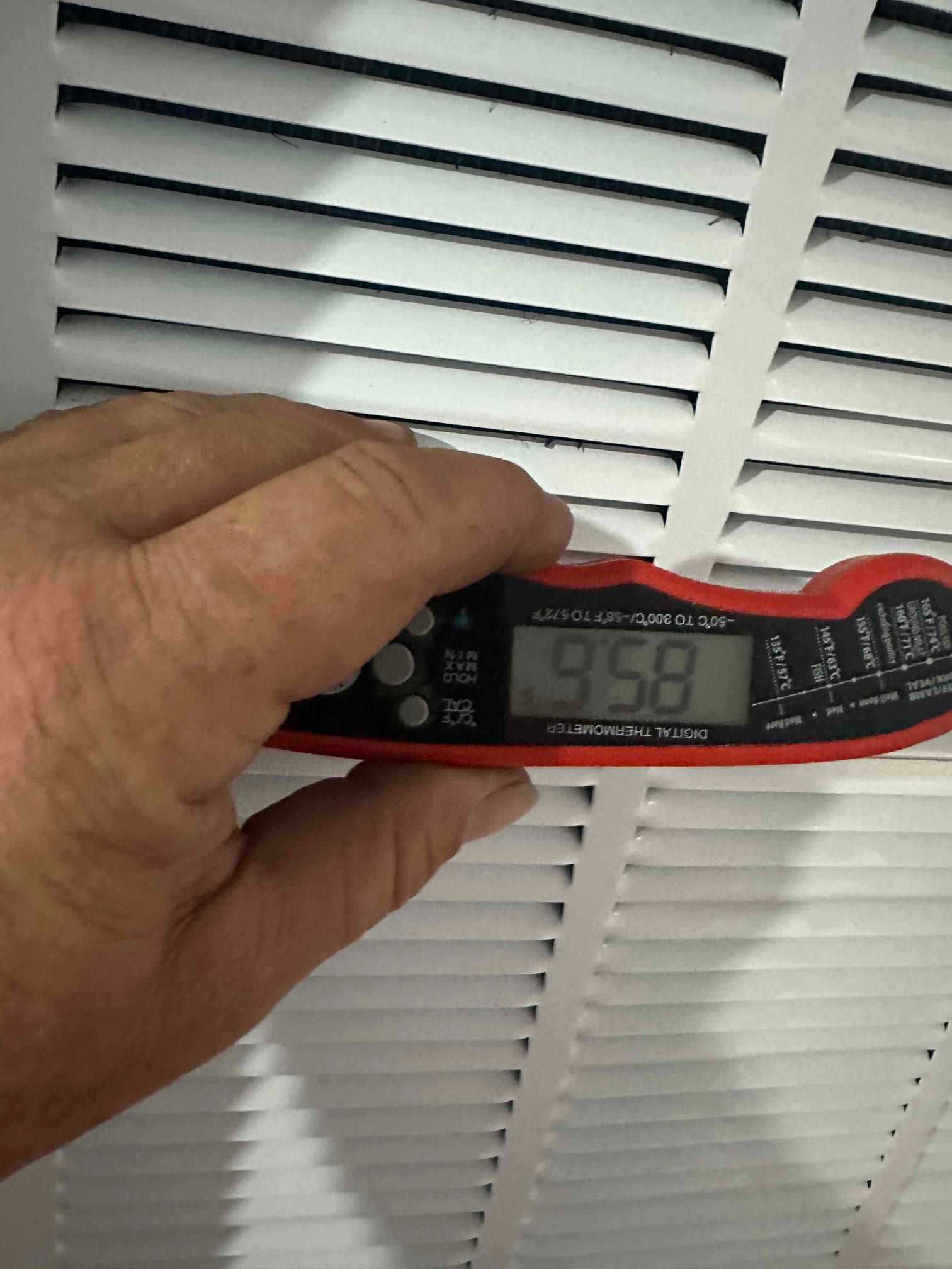 Digital thermometer held at a return grille shows 95.8 degrees Fahrenheit during air temperature check.