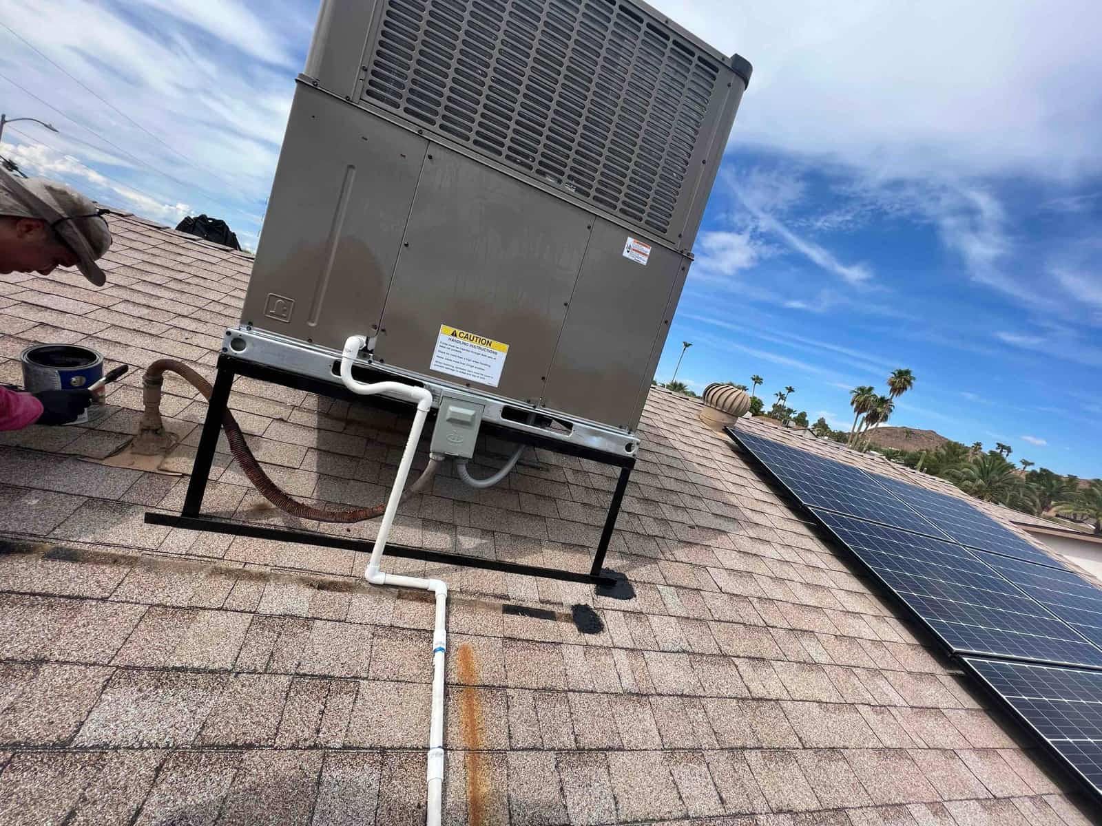 Rooftop package heat pump installed on stand with condensate drain, electrical whip, and sealed roof penetration beside solar panels.