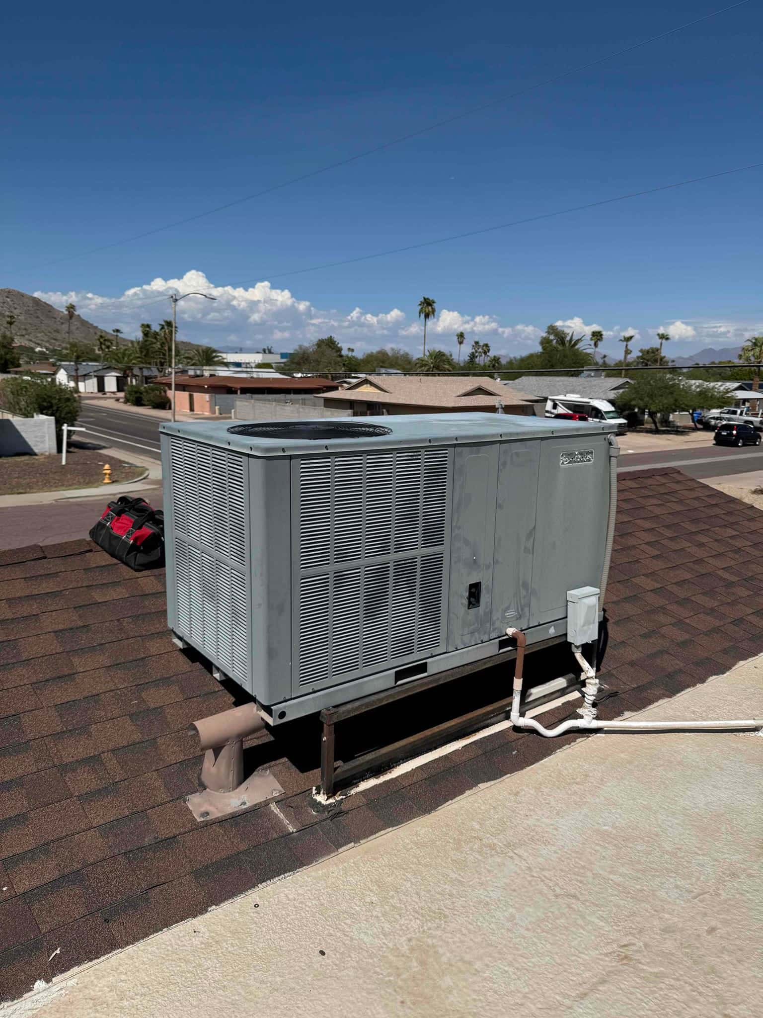 Rooftop packaged air conditioning unit on shingle roof, showing condenser fan section, service disconnect, and insulated refrigerant lines.