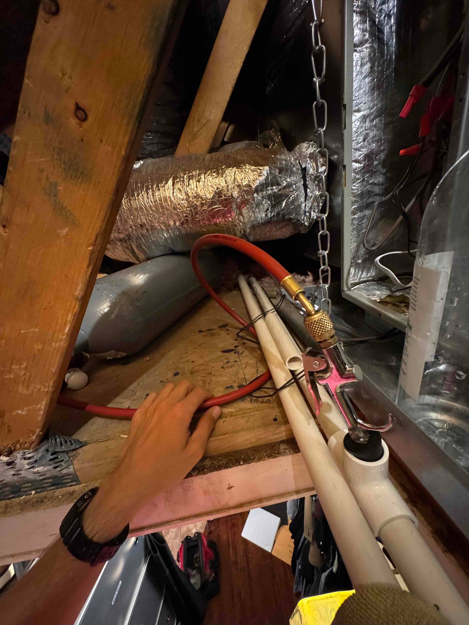 Technician’s hand accesses attic condensate drain lines near an air handler, showing PVC piping, cleanout, and insulated ductwork.