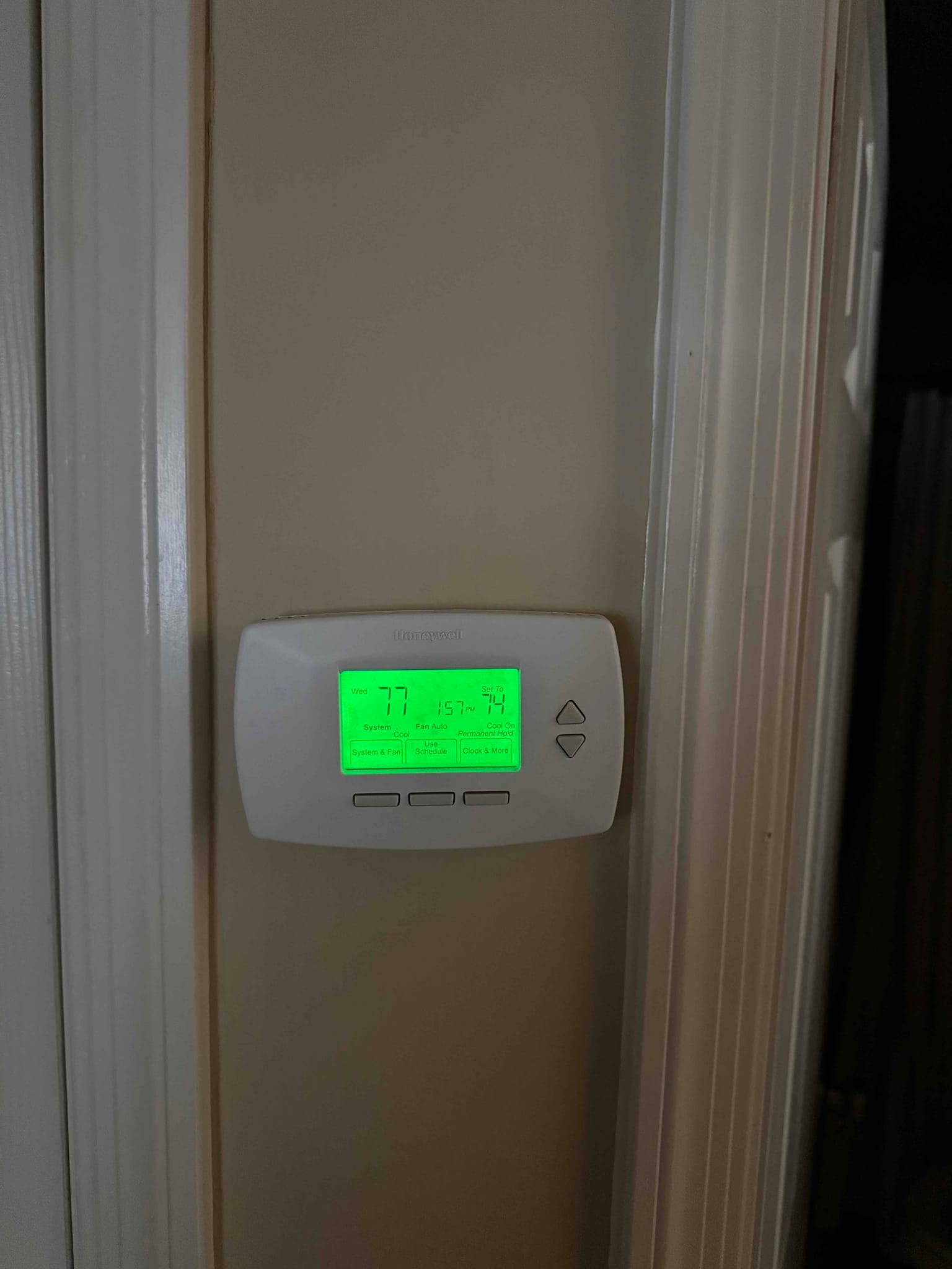Wall-mounted digital thermostat displays indoor temperature and setpoint with a green backlit screen and four control buttons.