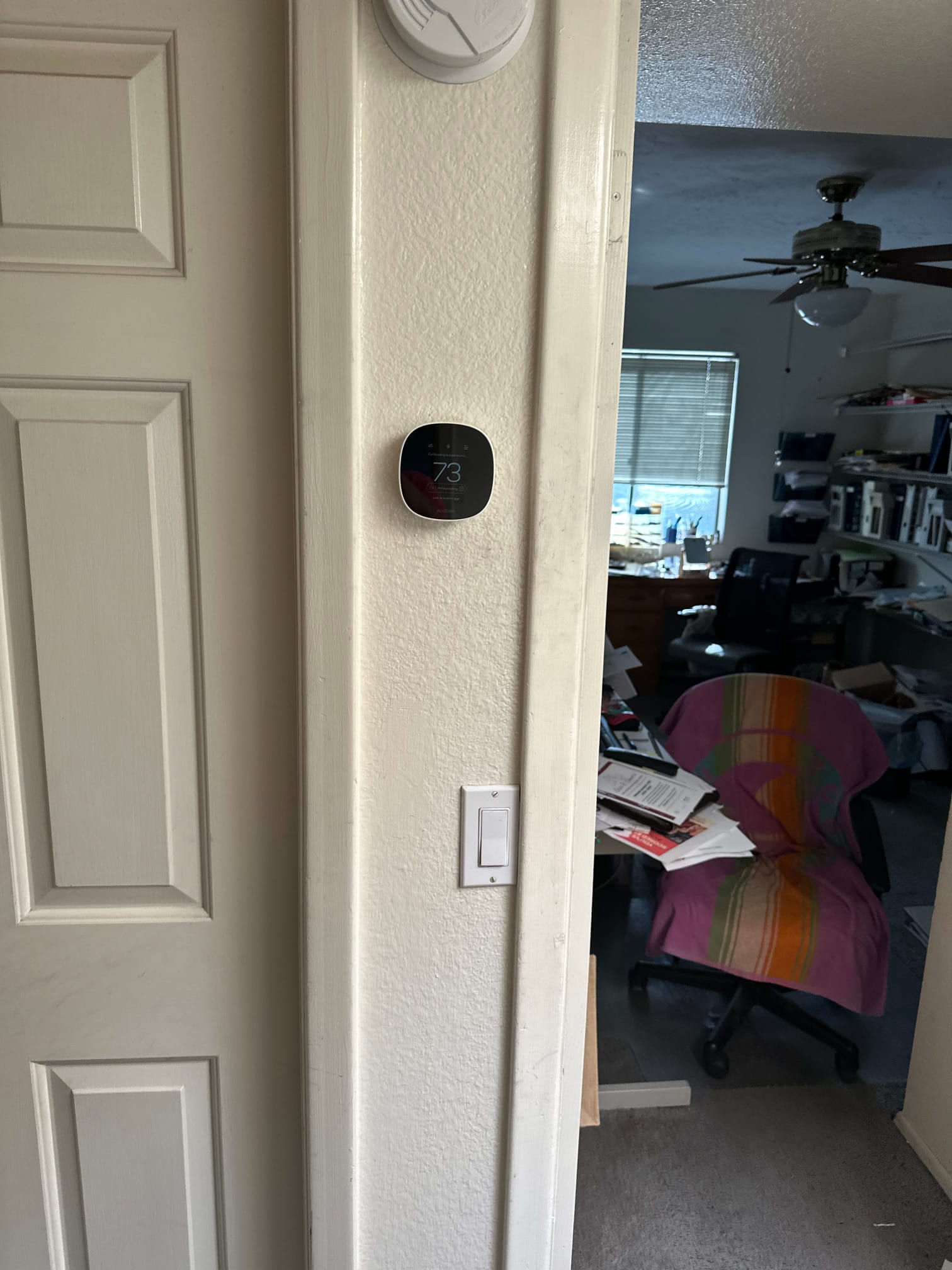 Wall-mounted smart thermostat installed and powered on near a smoke alarm, with adjacent room visible in the background.