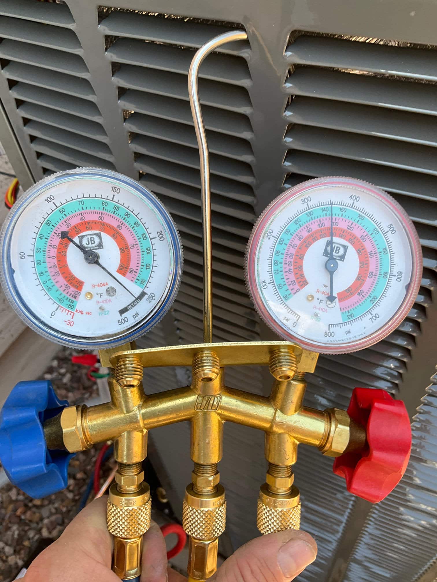 Analog manifold gauge set held in front of an outdoor condenser cabinet during installation, with hoses not yet connected.
