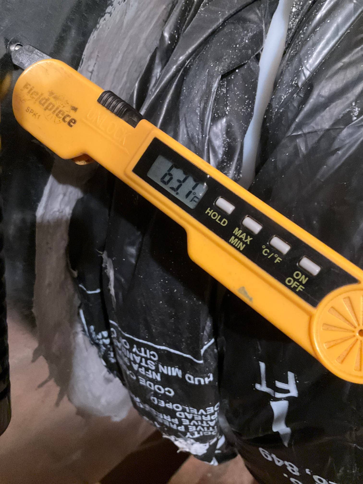 Digital thermometer shows 63.1°F while measuring air temperature near insulated ductwork during system checkout.